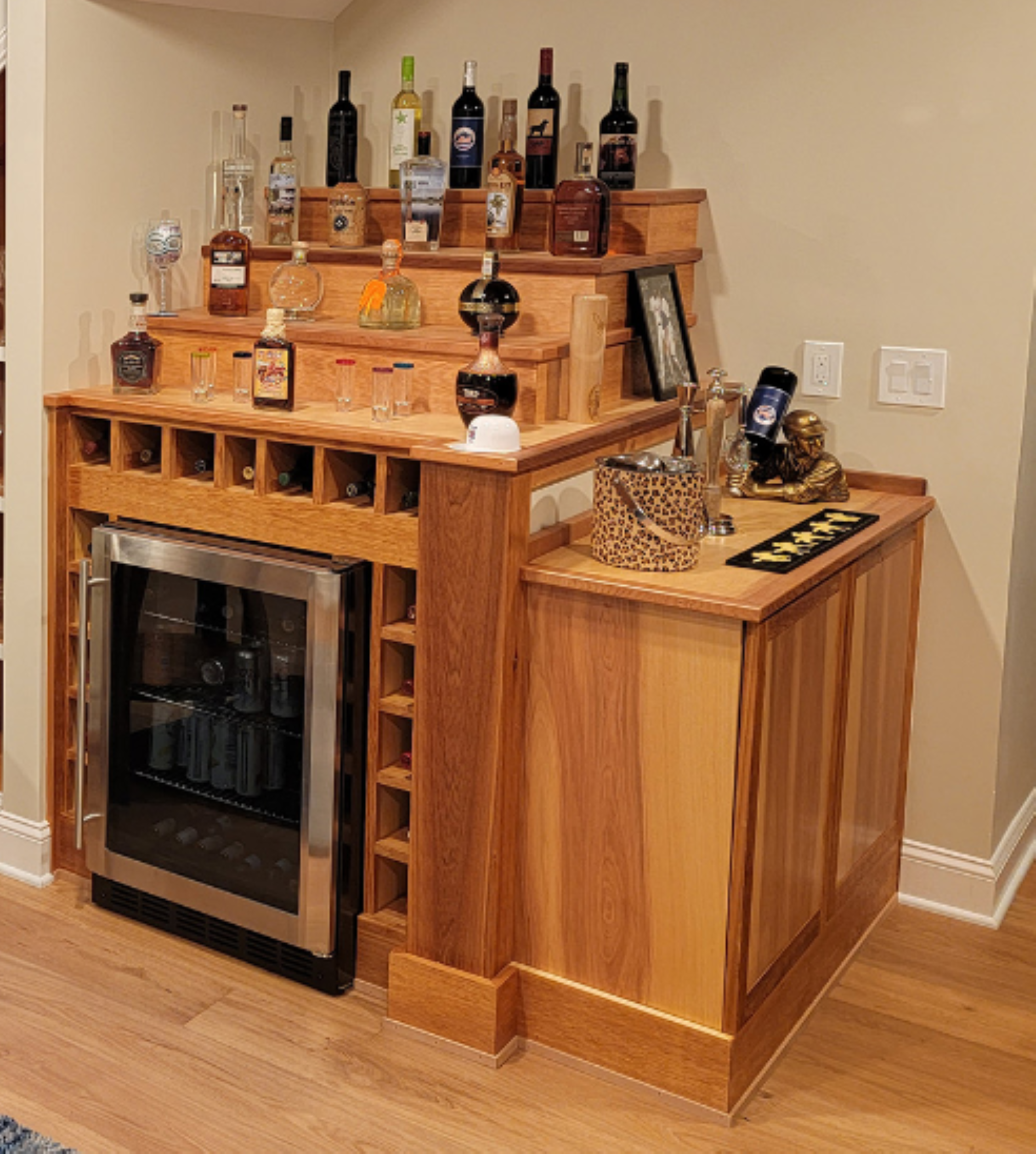 A wooden bar area with bottles of alcohol on tiered shelves, wine refrigerator