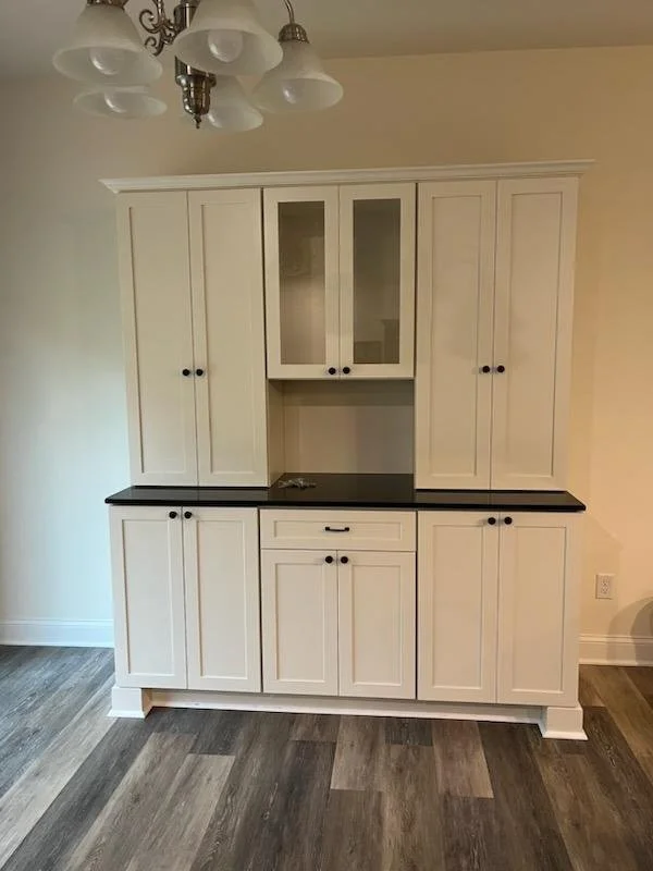 A kitchen cabinet with white doors and black handles, featuring glass-paneled upper doors, a black countertop, and wooden flooring.