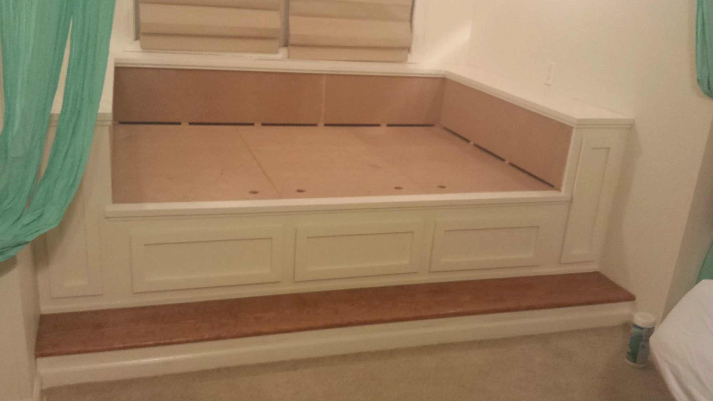 Image of a built-in platform bed frame with storage drawers underneath, made of white wood, with a column at the end, in a bedroom with beige carpet and a bottle of medicine or supplement on the floor