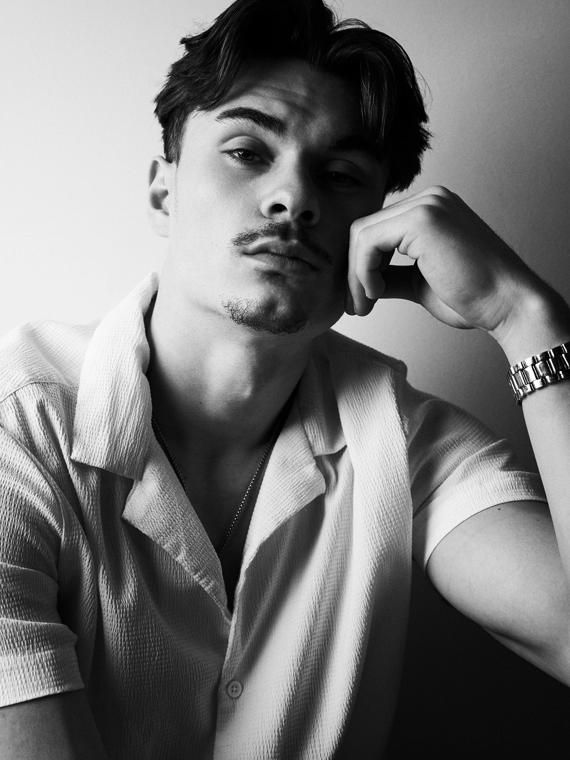 Black and white photo of a young man with dark hair, a mustache and goatee, wearing a short-sleeved, collared shirt and a wristwatch, looking at the camera with a relaxed expression, his hand resting on his chin.