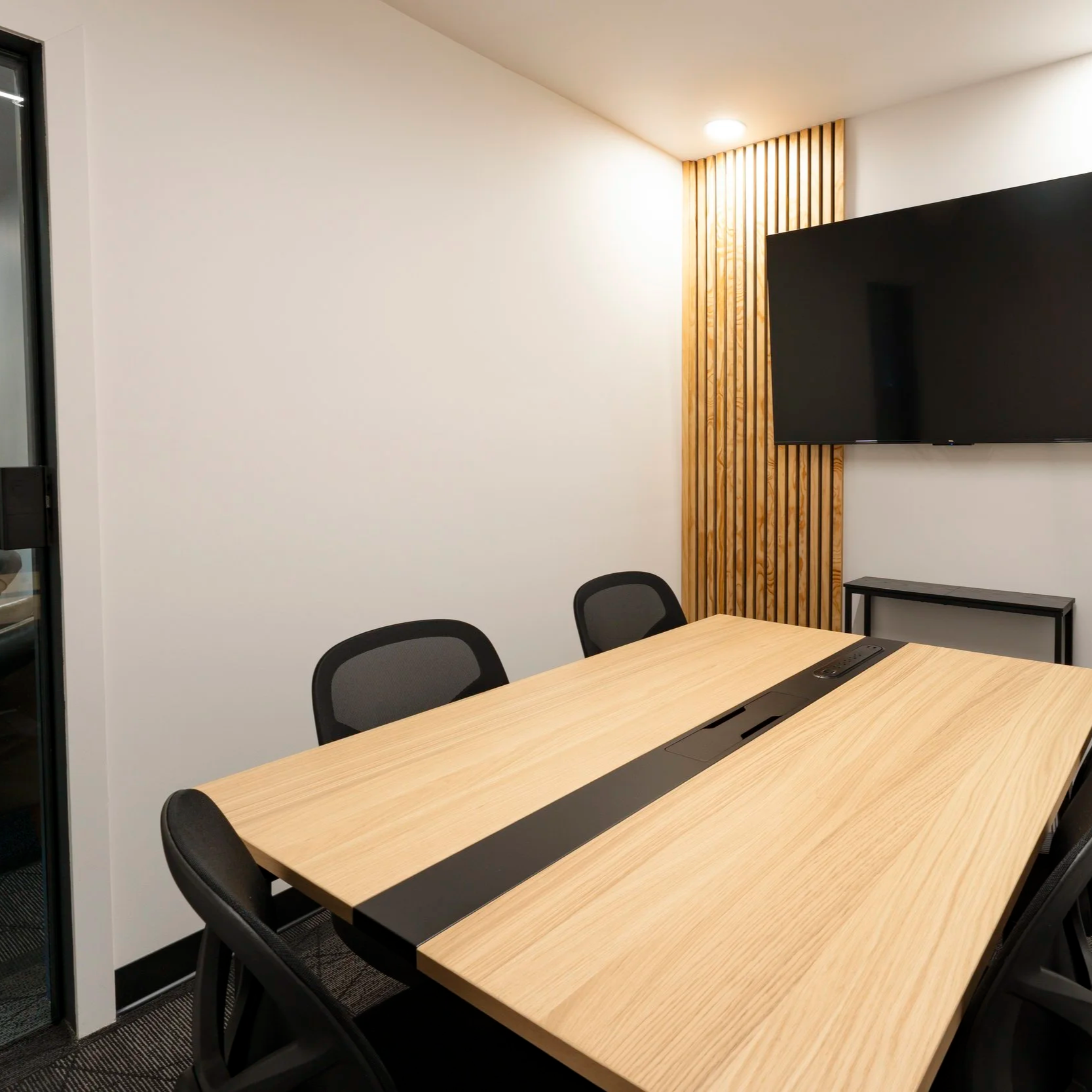 A modern conference room with a wooden table, black chairs, a wall-mounted flat-screen TV, and a decorative wood panel on the wall.