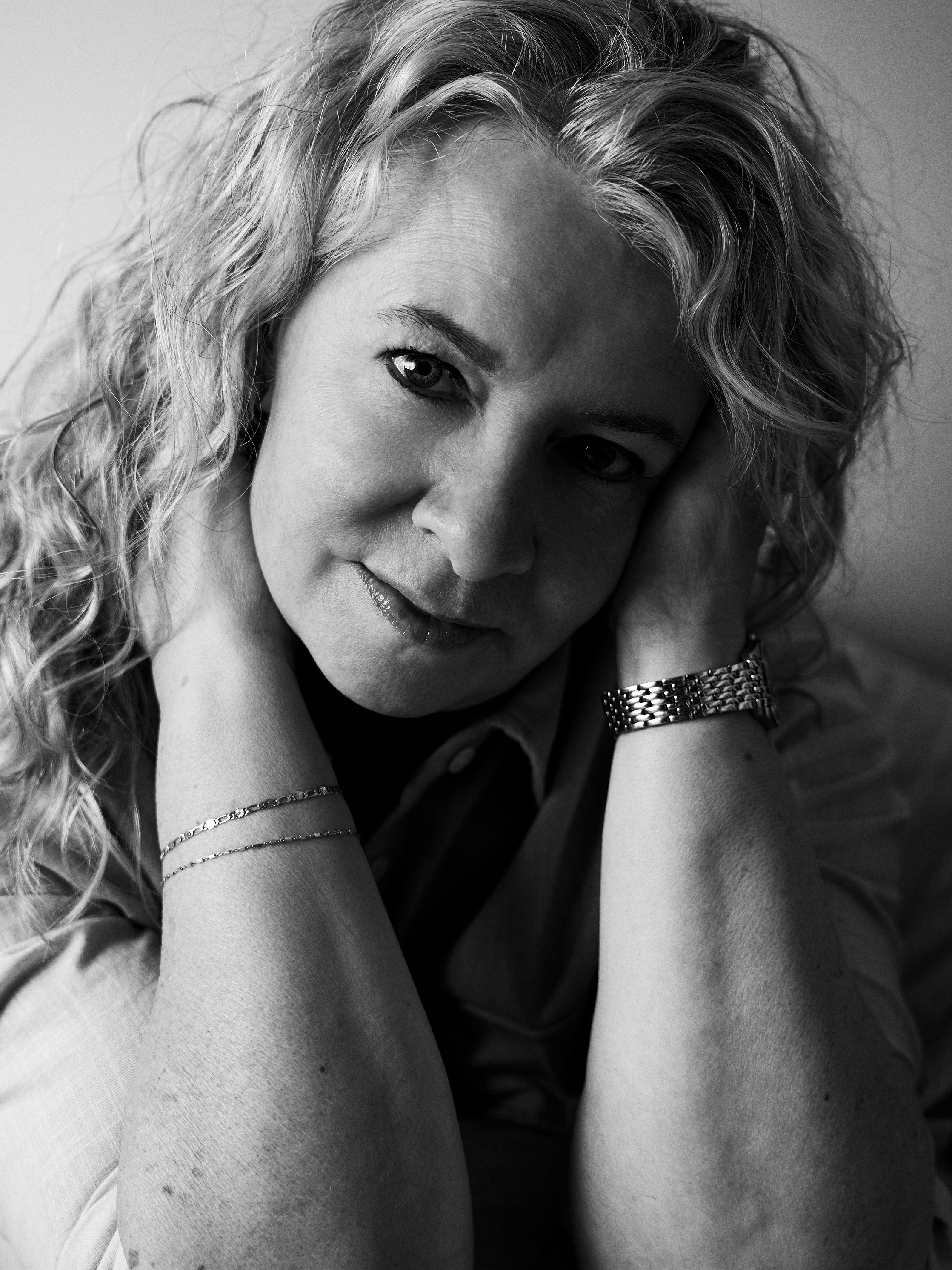 A black and white photograph of a woman with curly hair resting her head on her arms, looking softly at the camera.