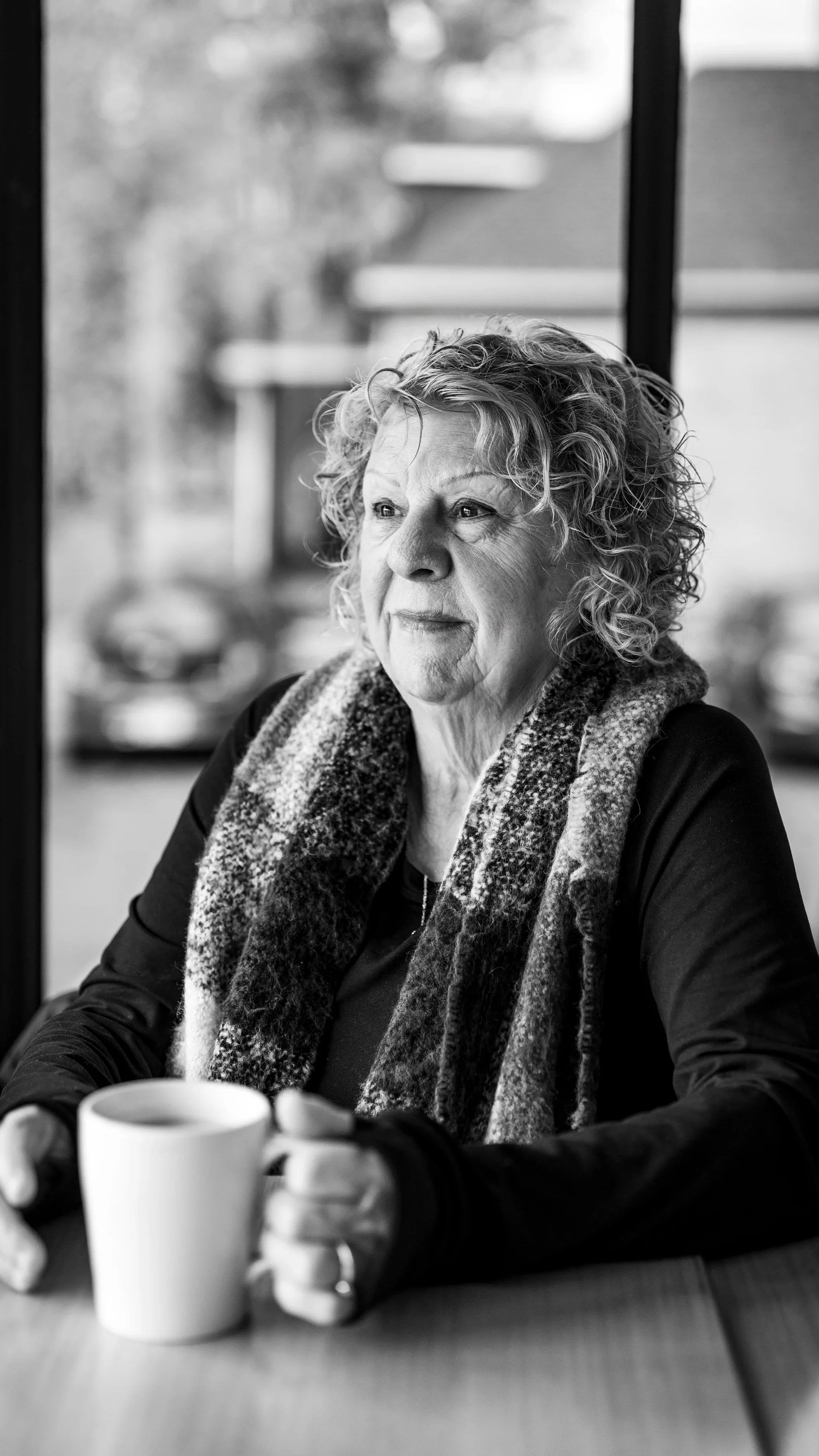 An elderly woman with curly hair sits at a table holding a mug, gazing thoughtfully out a window, in a black and white photo.