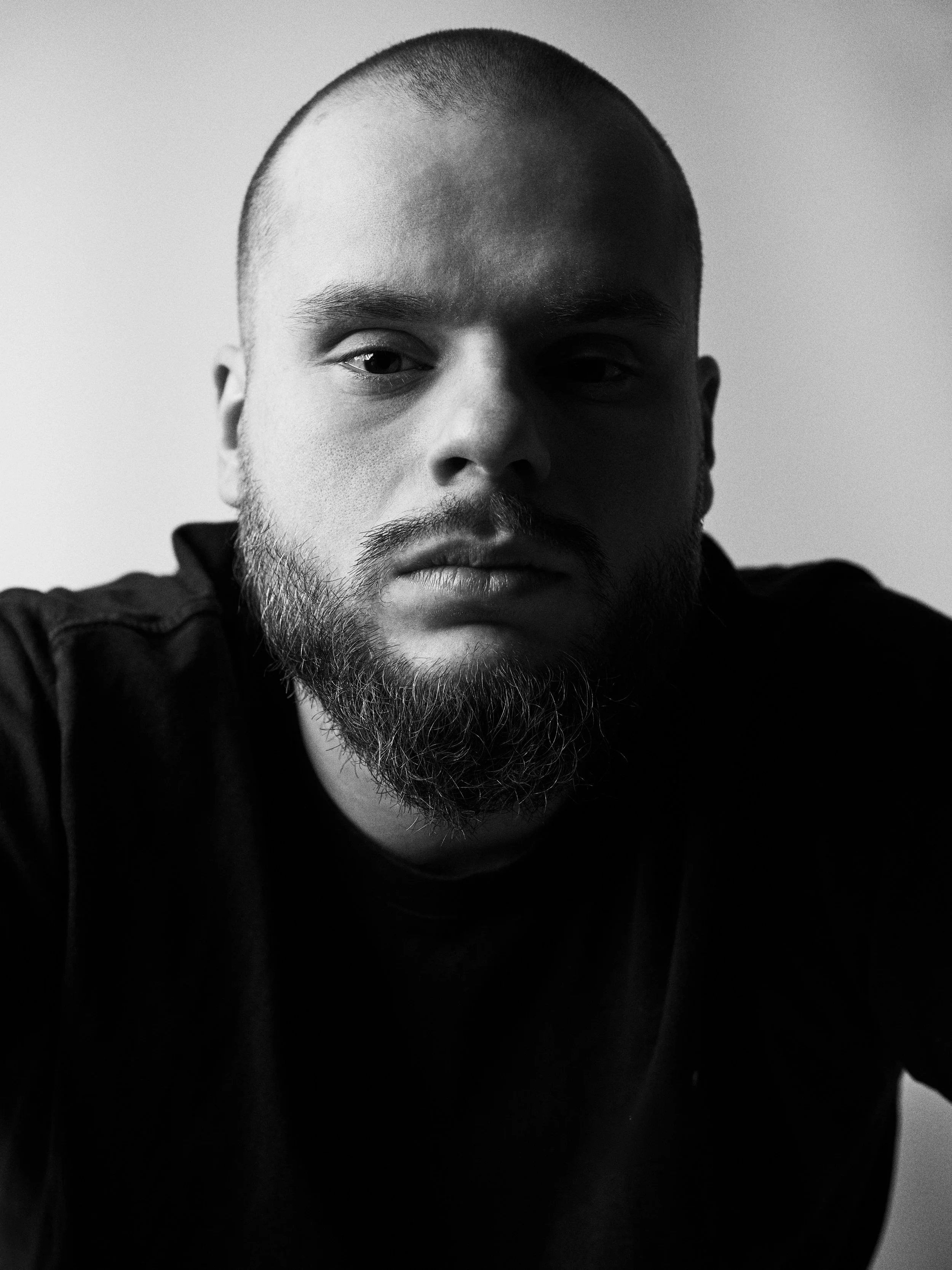 Close-up black and white photo of a man with a shaved head and a beard, wearing a dark shirt, looking into the camera with a neutral expression.