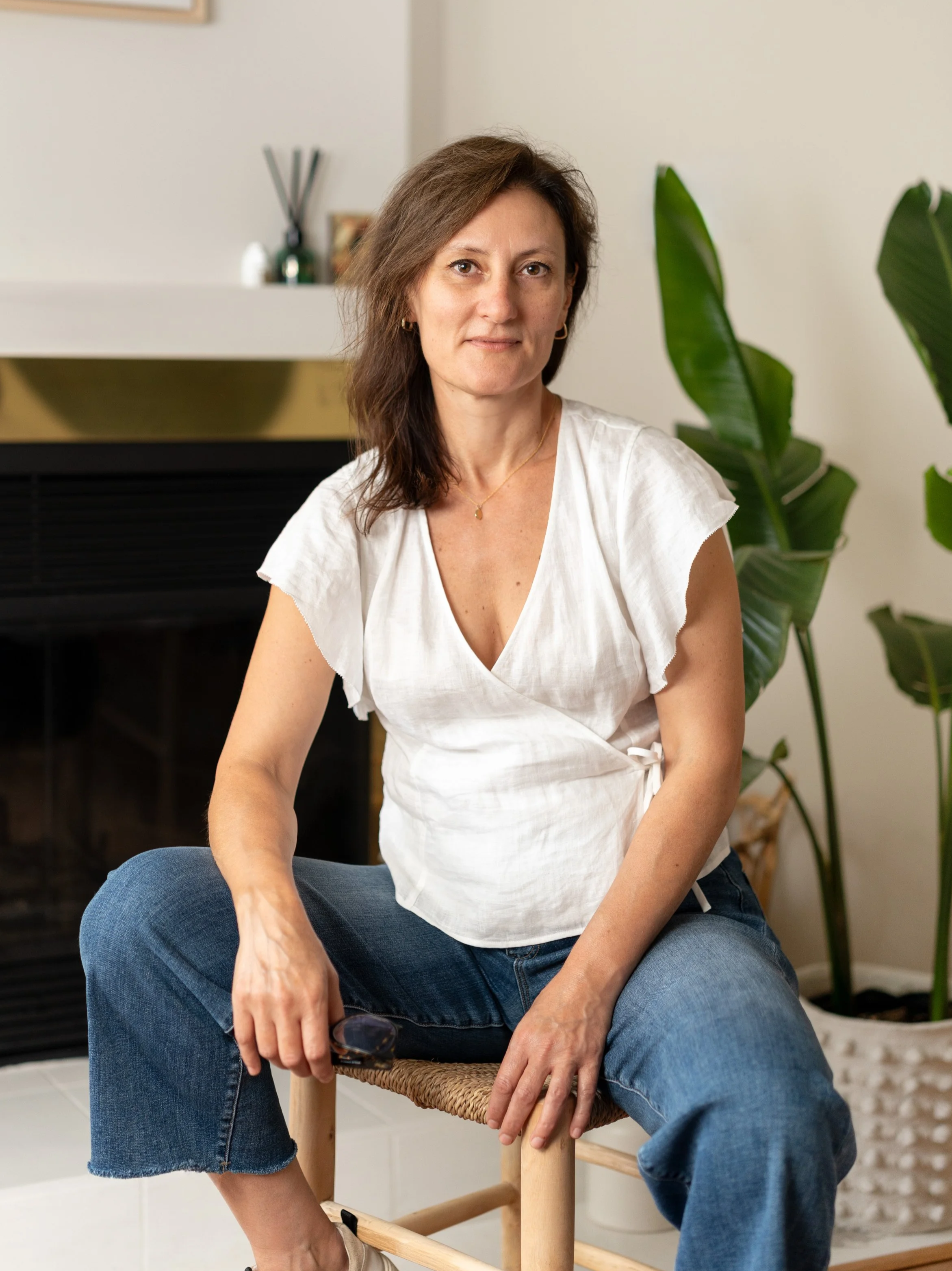 A woman with brown hair, wearing a white top and blue jeans, sitting on a woven chair in a room with a fireplace and large green houseplants.