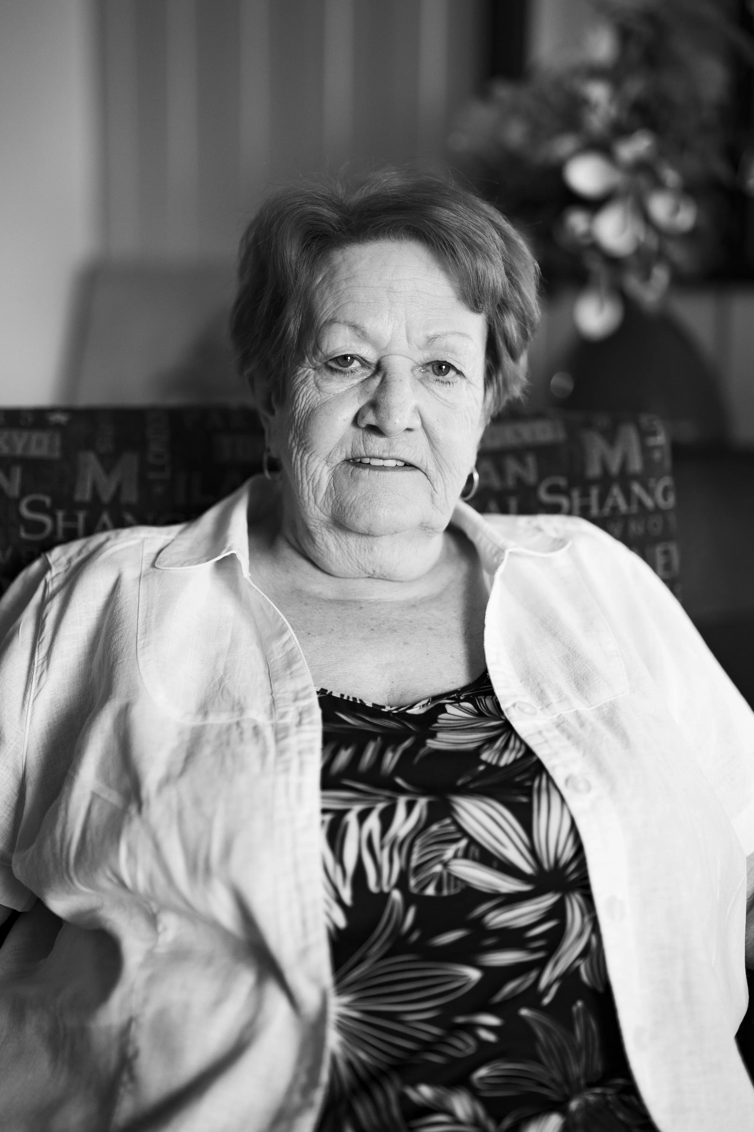 Black and white photo of an older woman with short hair, wearing a light-colored shirt, sitting indoors with a blurred background, including a plant, with a serious expression on her face.