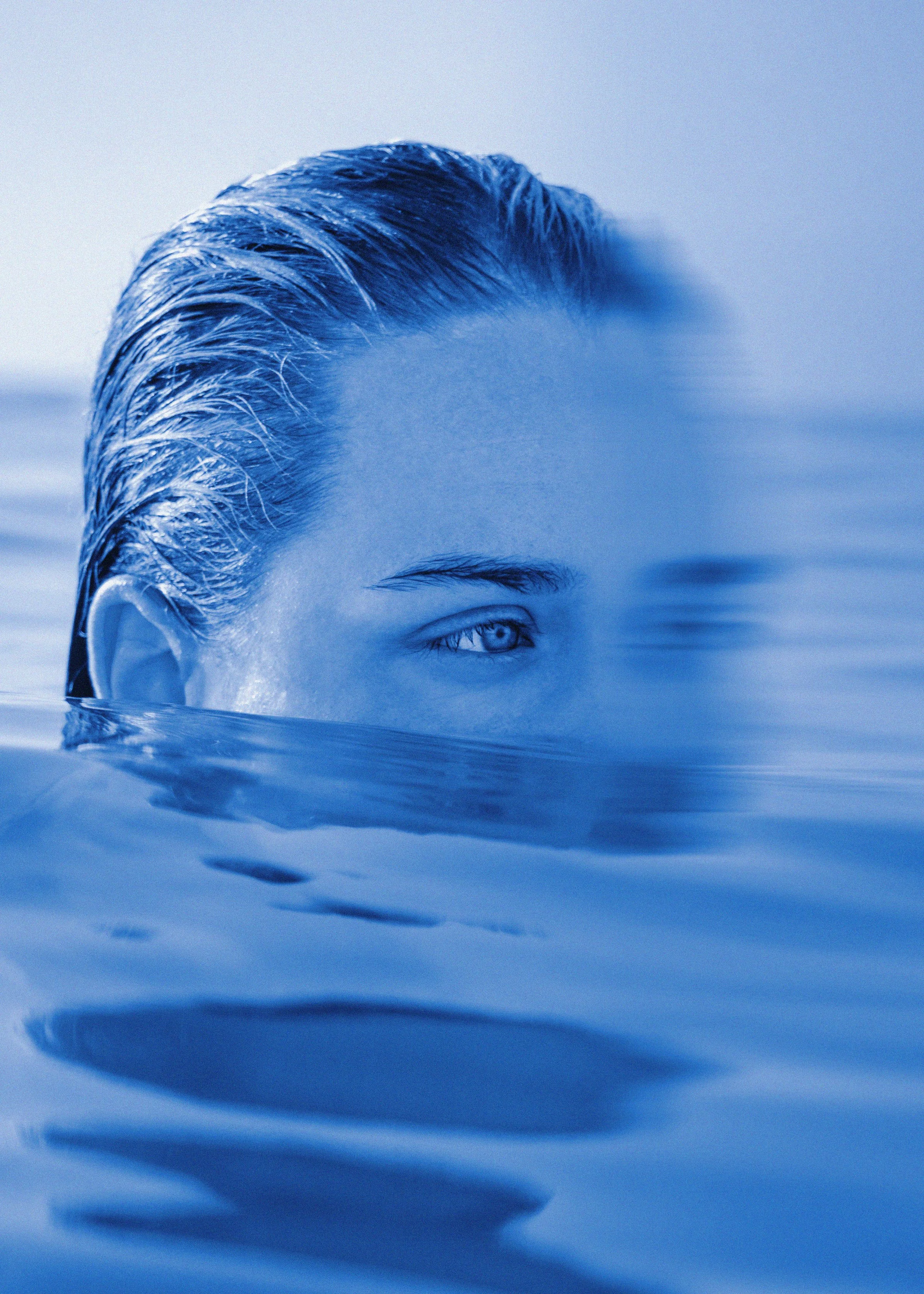 Close-up of a person's face and hair partially submerged in water, with only the eye, part of the forehead, and hair visible above the waterline, creating a serene and mysterious atmosphere.