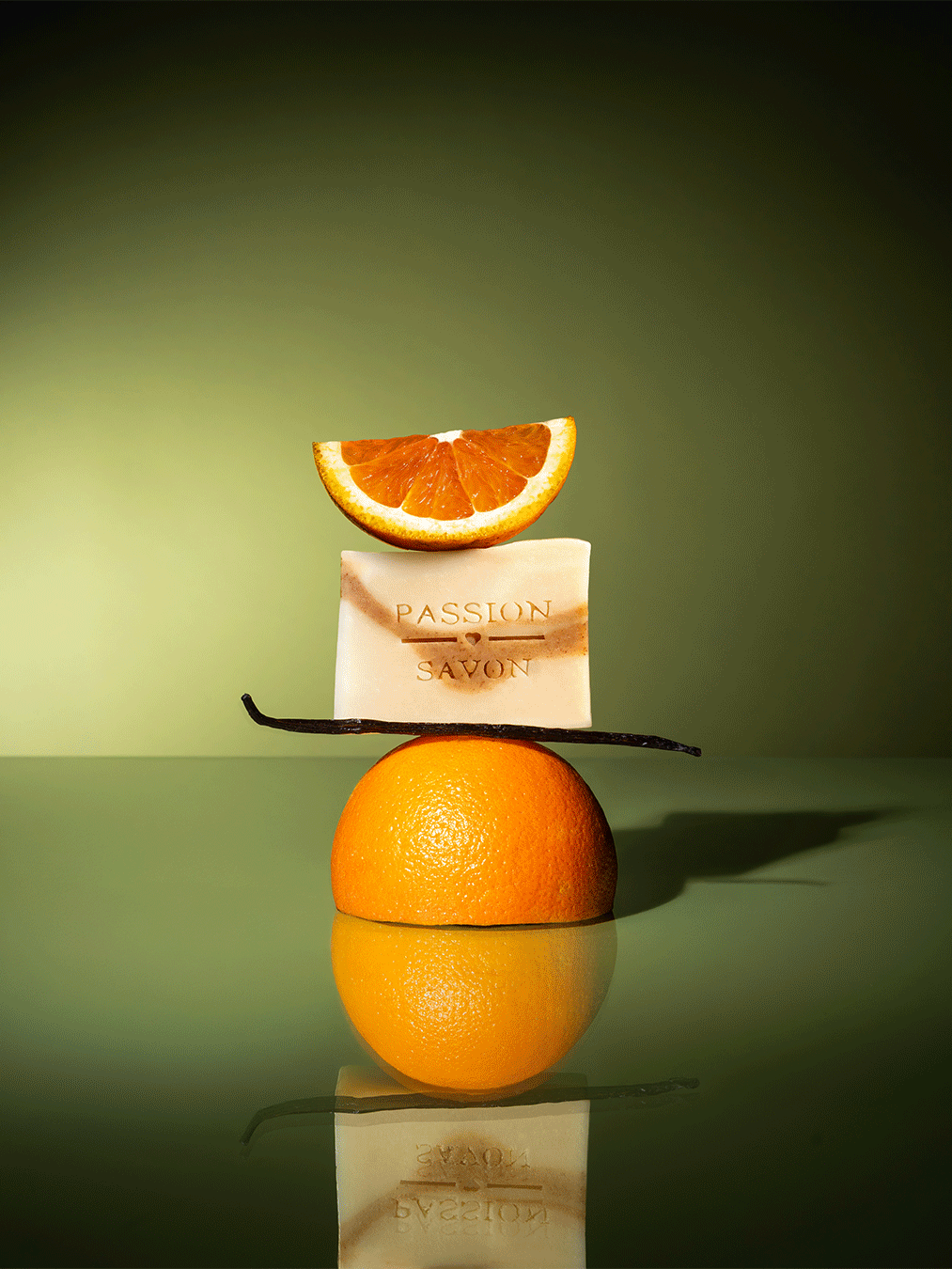 A soap bar labeled 'Passion Savon' with a slice of orange on top, an orange half balanced on the soap, and a vanilla bean, all arranged on an orange half, with a green background.