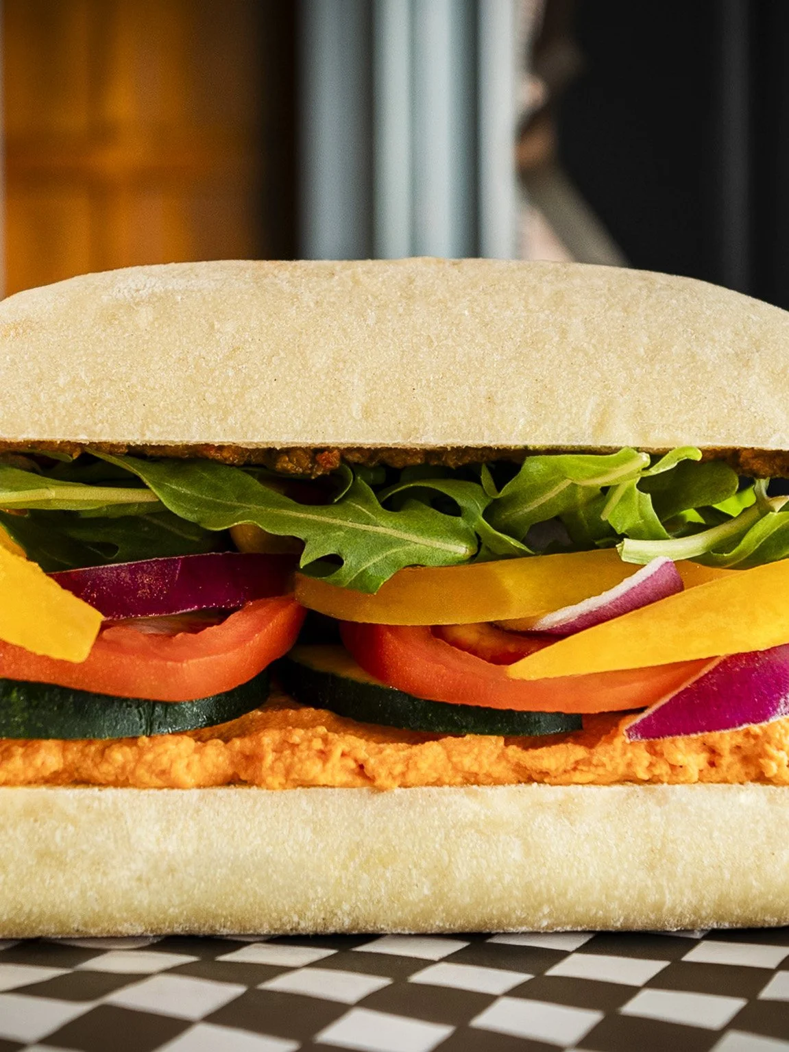 Close-up of a submarine sandwich with bread, lettuce, tomato, cucumber, radish, red onion, and a layer of orange spread.