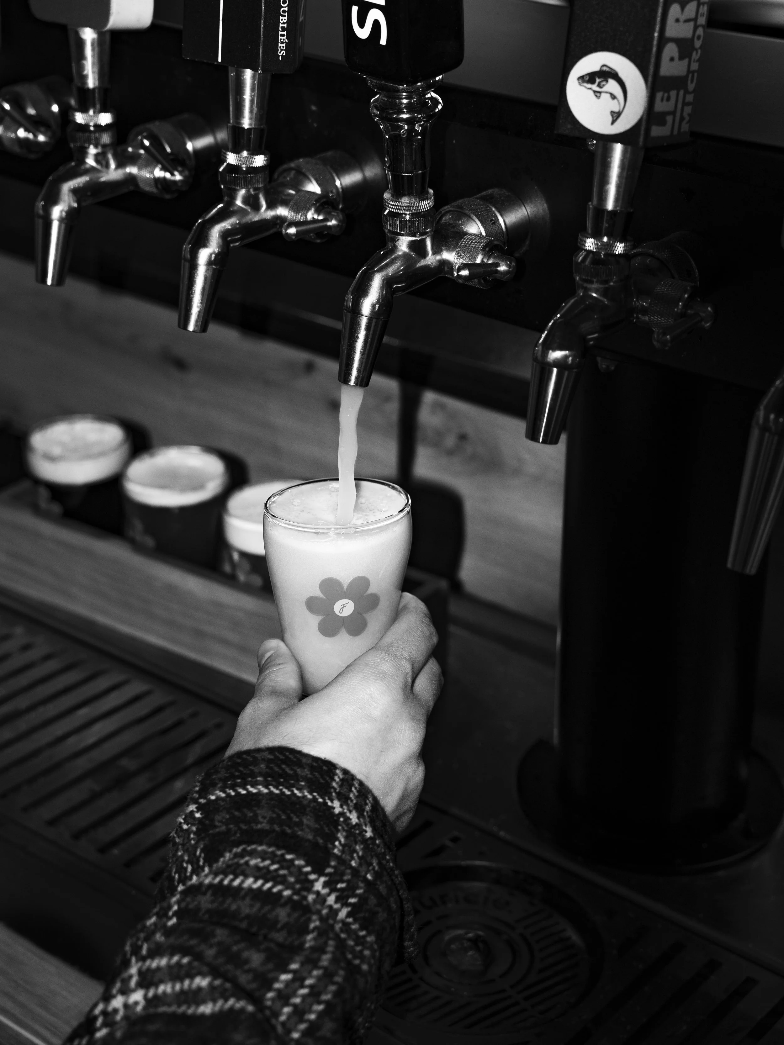 A person, dressed in a patterned sweater, holding a glass under a tap with beer or liquid pouring into it. The tap appears to be part of a bar or pub setting.