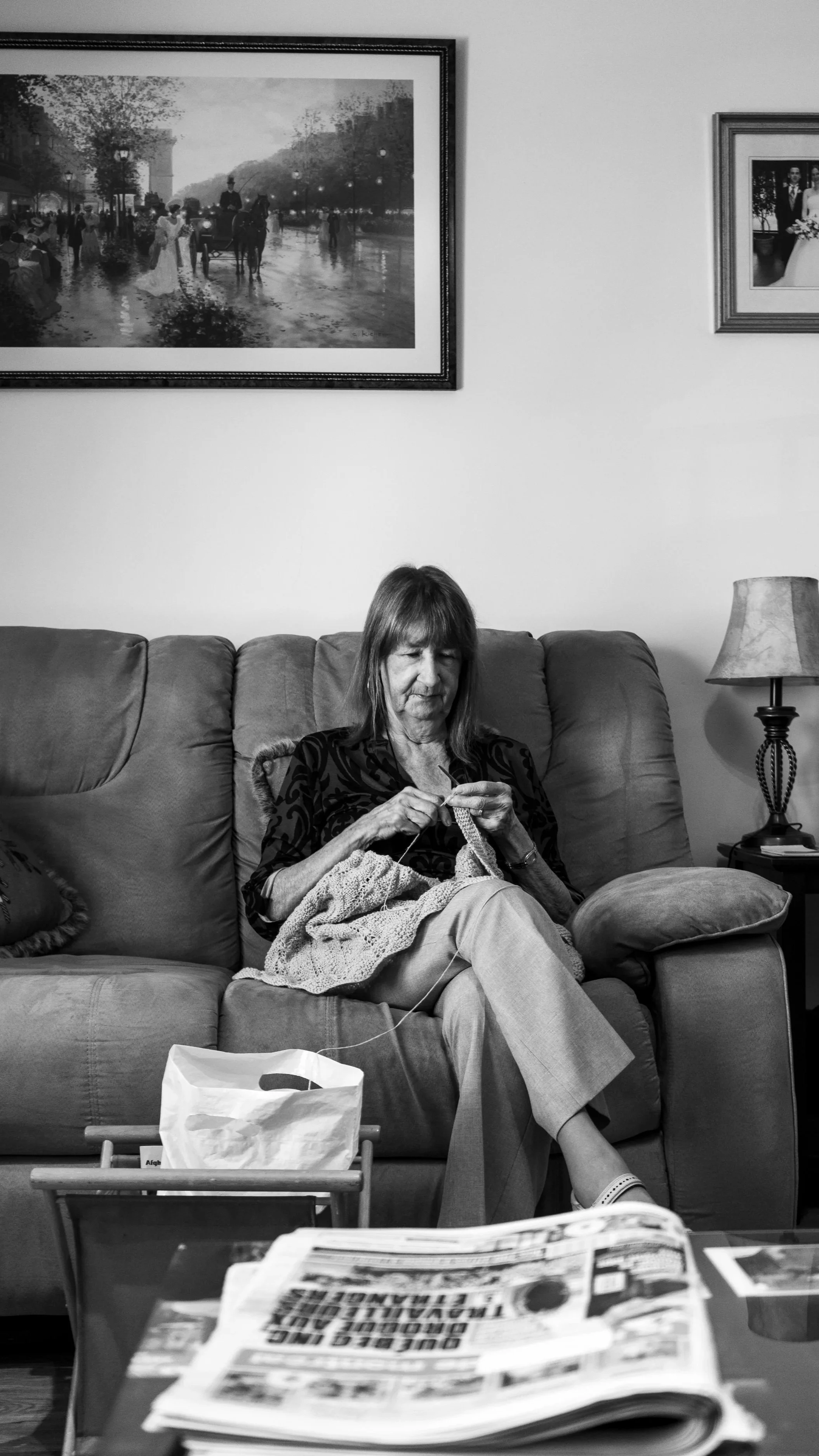 A woman knitting on a sofa in a living room. There are framed pictures hanging on the wall behind her, a table lamp to her right, and a newspaper and paper bag in front of her.