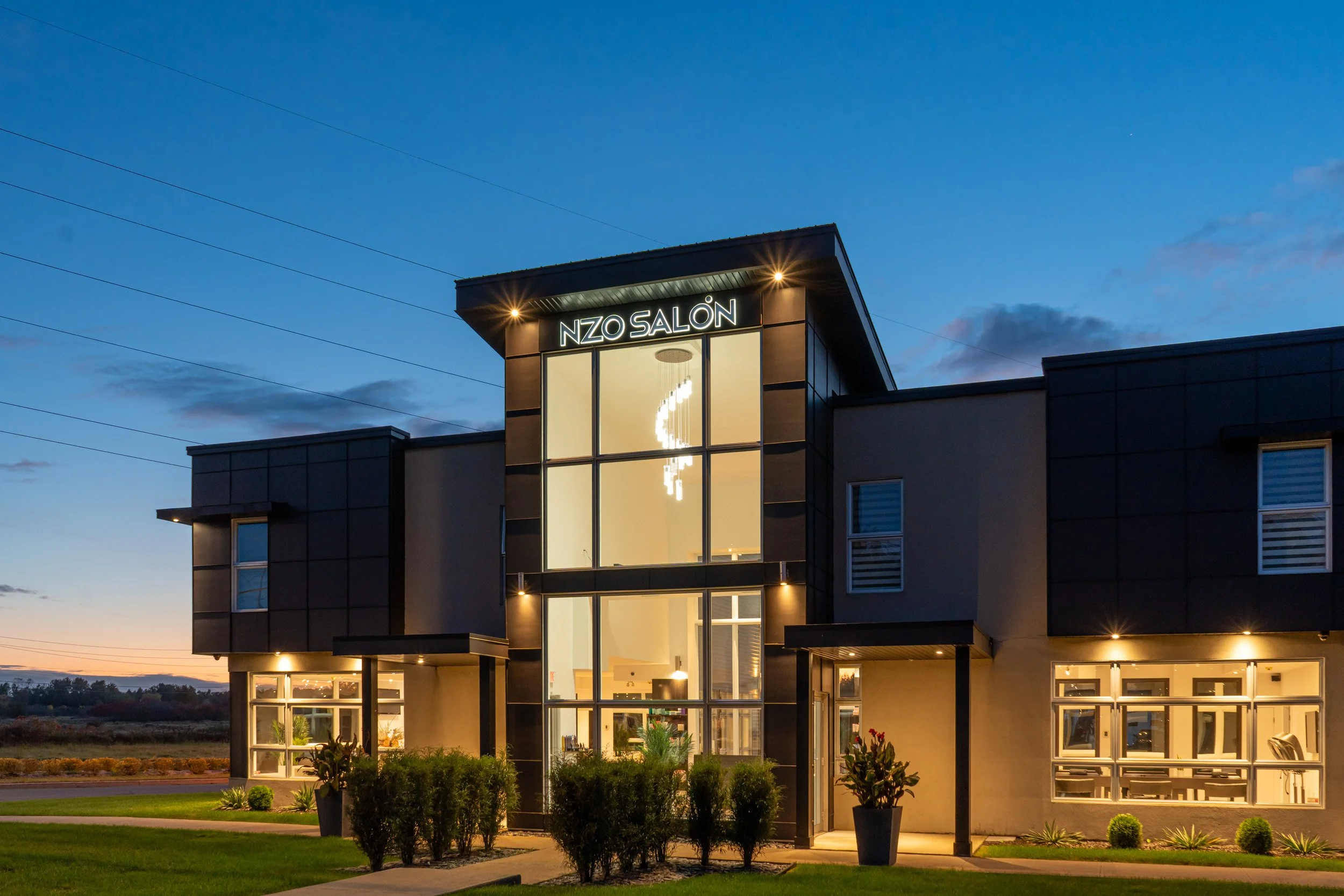 Modern commercial building with large glass windows and neon sign that reads 'NZO SALÓN' at dusk. Exterior lighting highlights the structure, and there are landscaped bushes and a lawn in front.