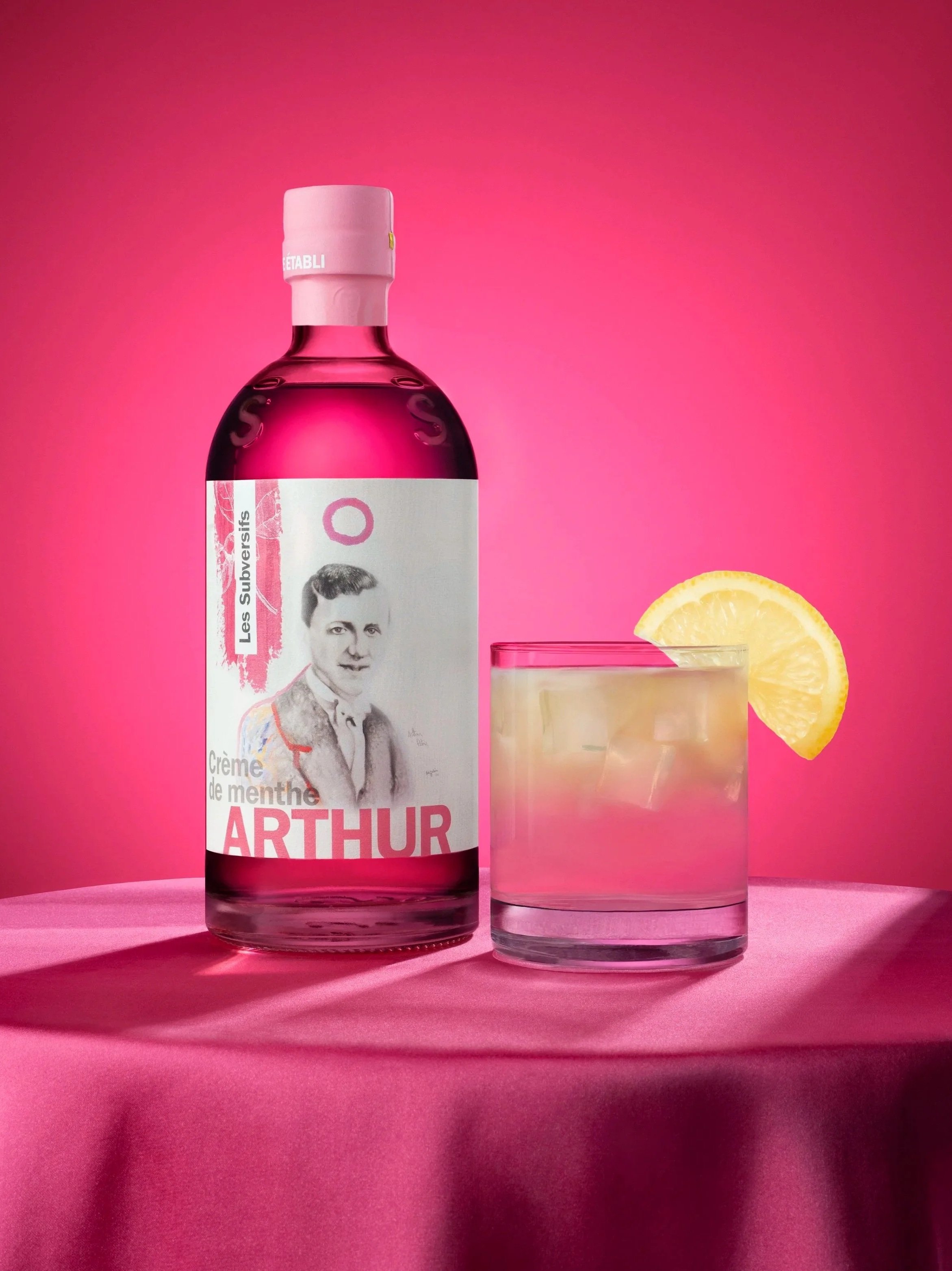 A bottle of Menthe Arthur herbal liqueur with a floral label, placed on a pink surface with a glass of water and lemon wedges nearby, against a pink background.