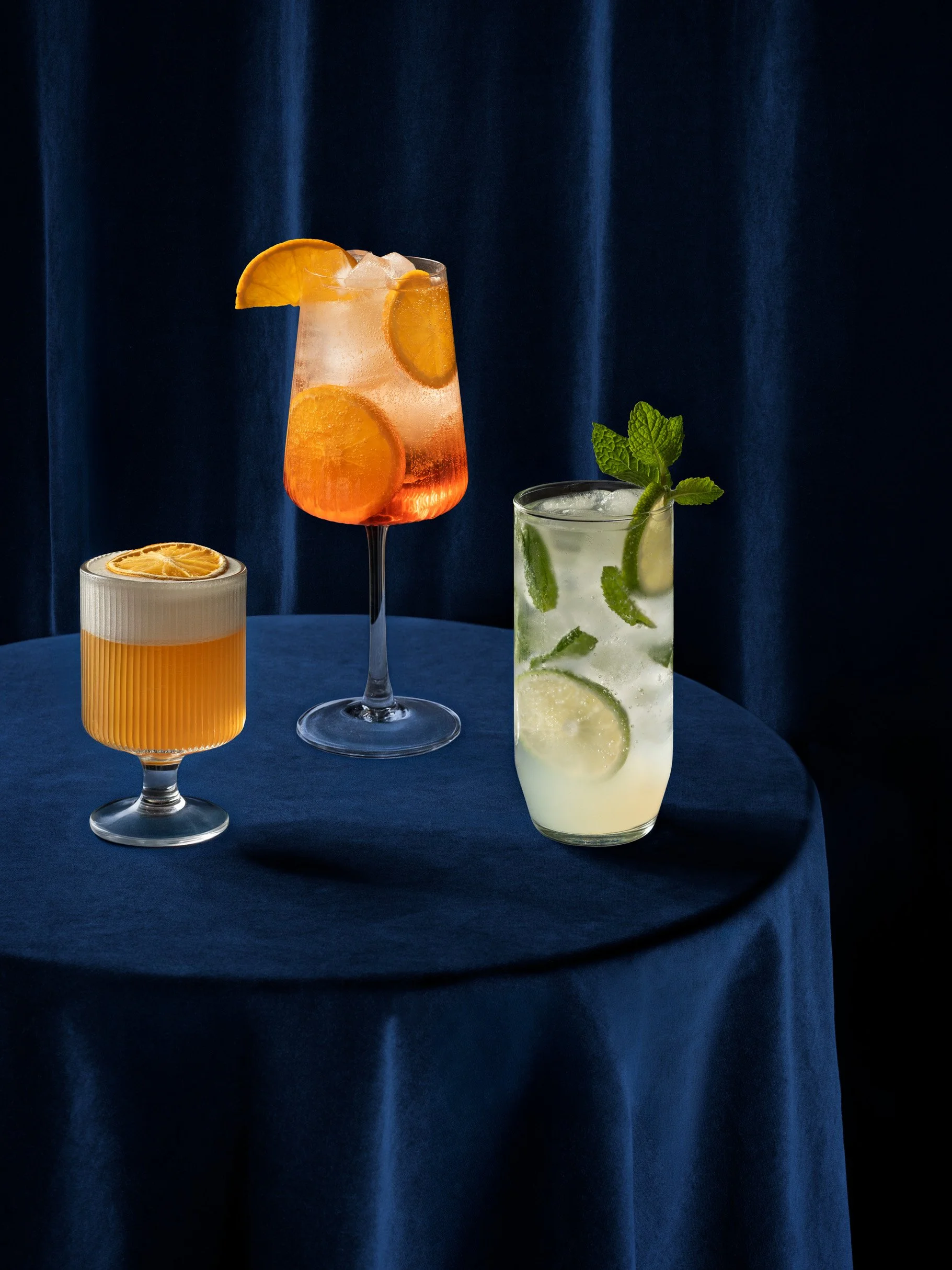 Three colorful beverages: a cocktail with orange slices and ice in a tall champagne flute, a yellow drink with a lemon slice in a ribbed glass, and a mojito with mint, lime, and ice in a highball glass