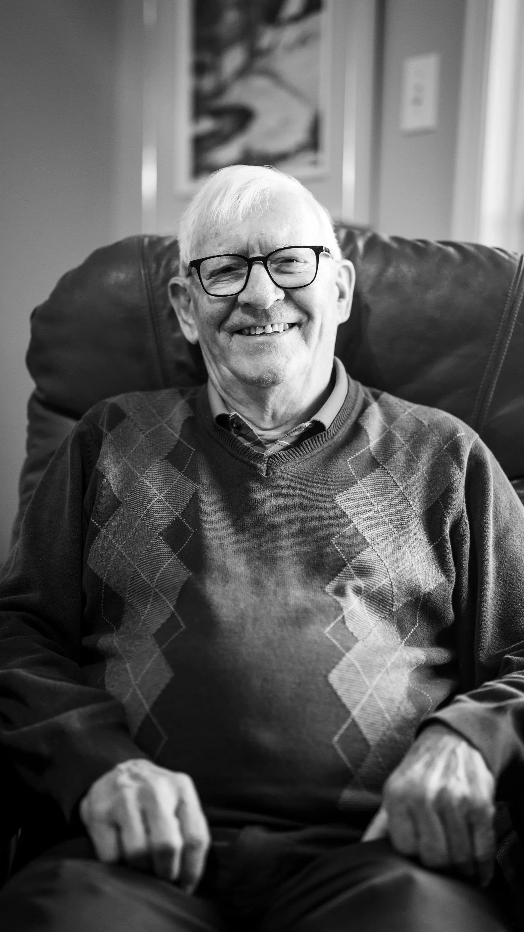 A smiling elderly man with glasses sitting in a leather chair, wearing a sweater with an argyle pattern, in a cozy room with a painting on the wall.