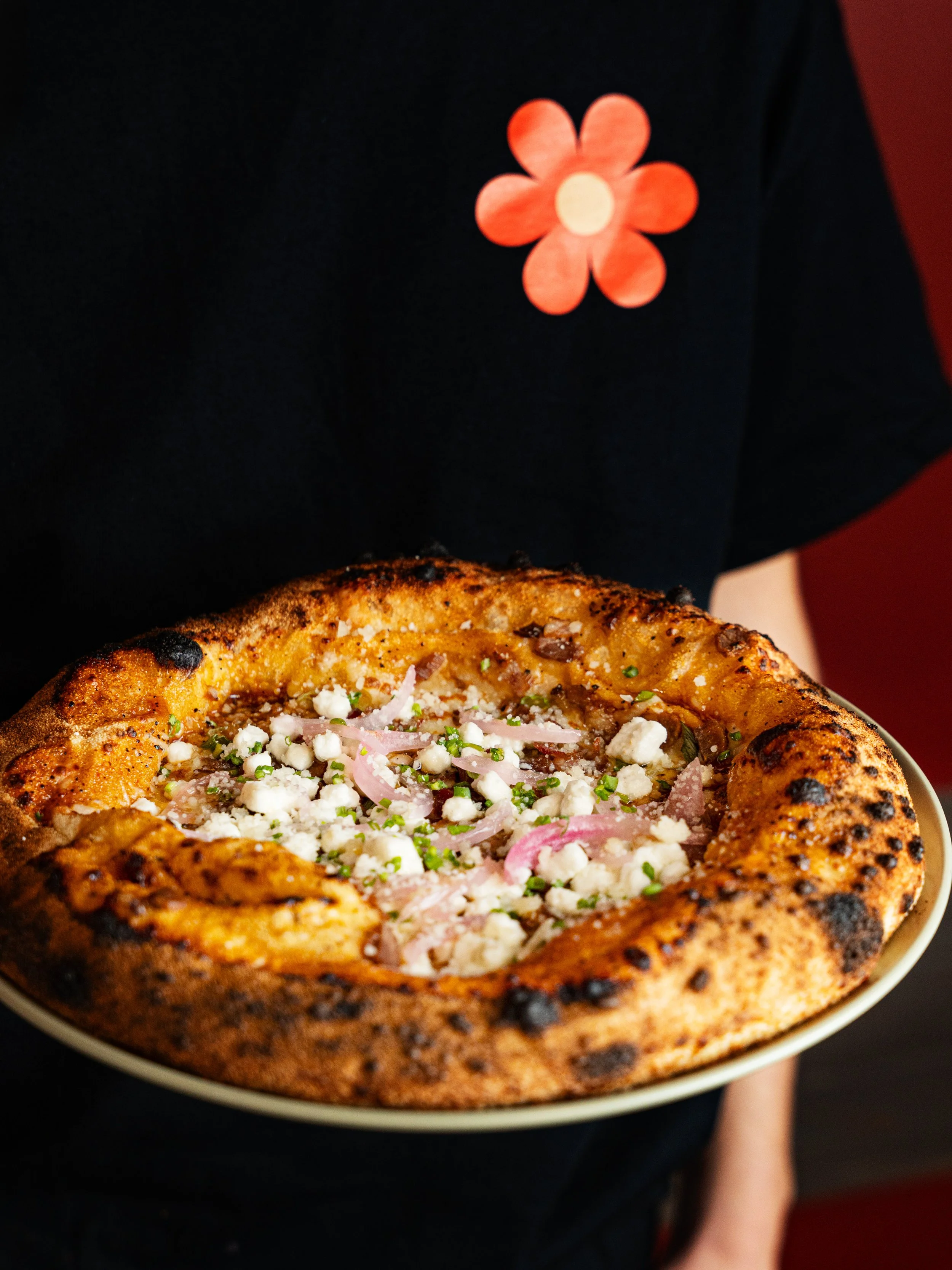 Close-up of a pizza with a golden, slightly charred crust, topped with crumbled white cheese, thin slices of onion, small green herbs, and possibly pieces of ham or bacon.