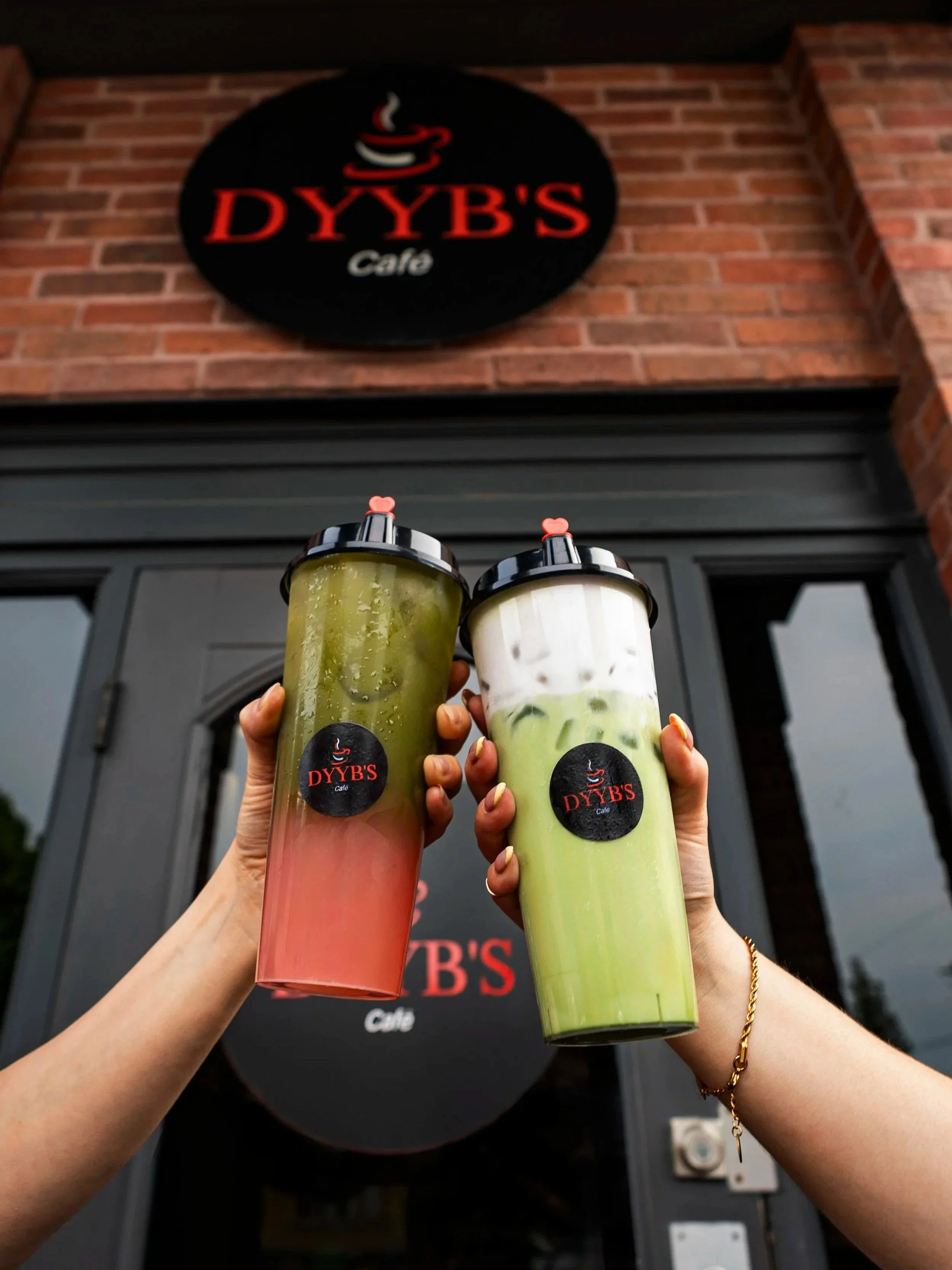 Two hands holding large plastic cups with beverages in front of Dyby's Cafe sign, which is mounted on a brick wall.