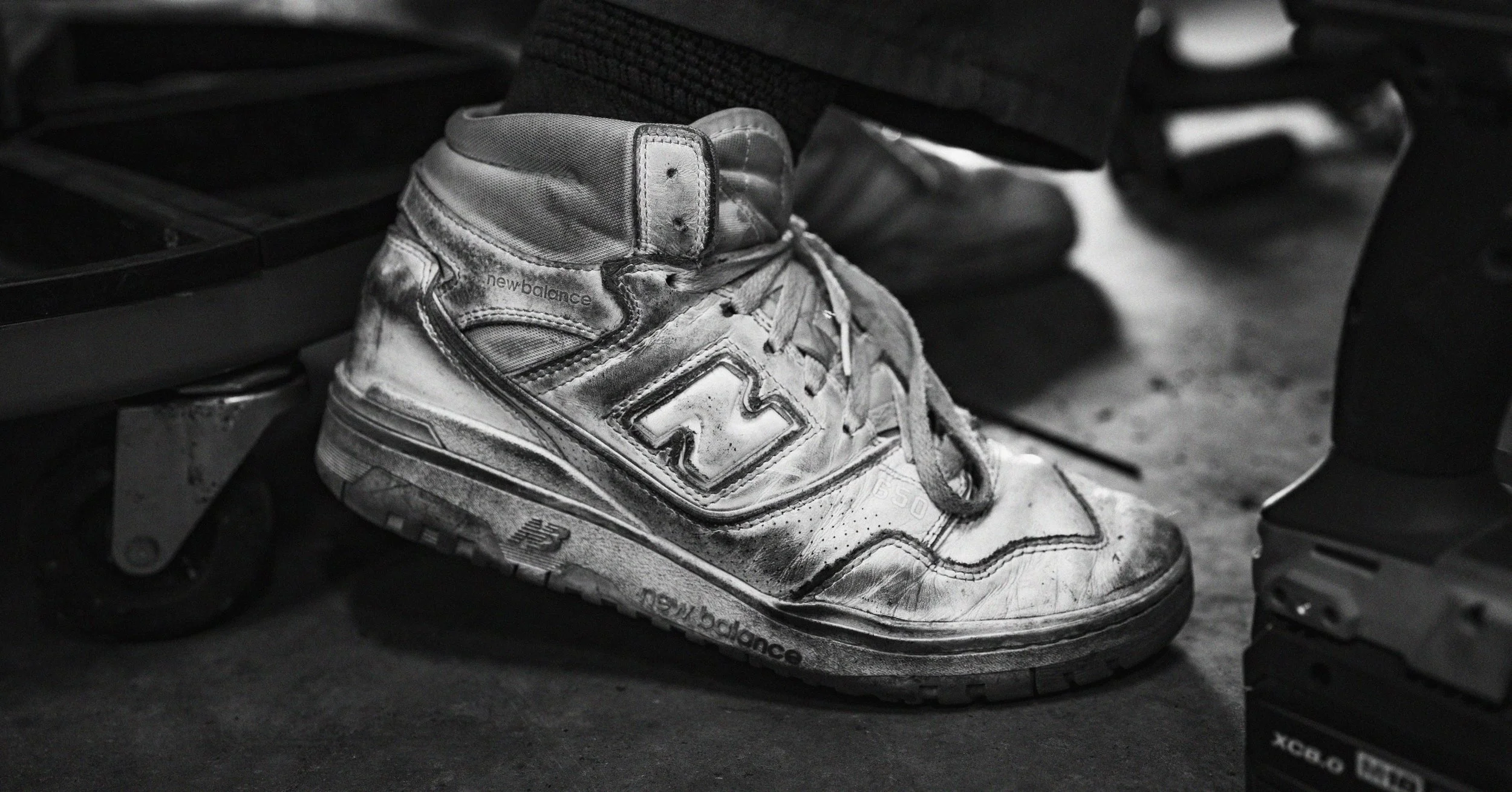 A worn and dirty white and gray New Balance sneaker on a gym bench, with black pants and the edge of another shoe visible nearby.
