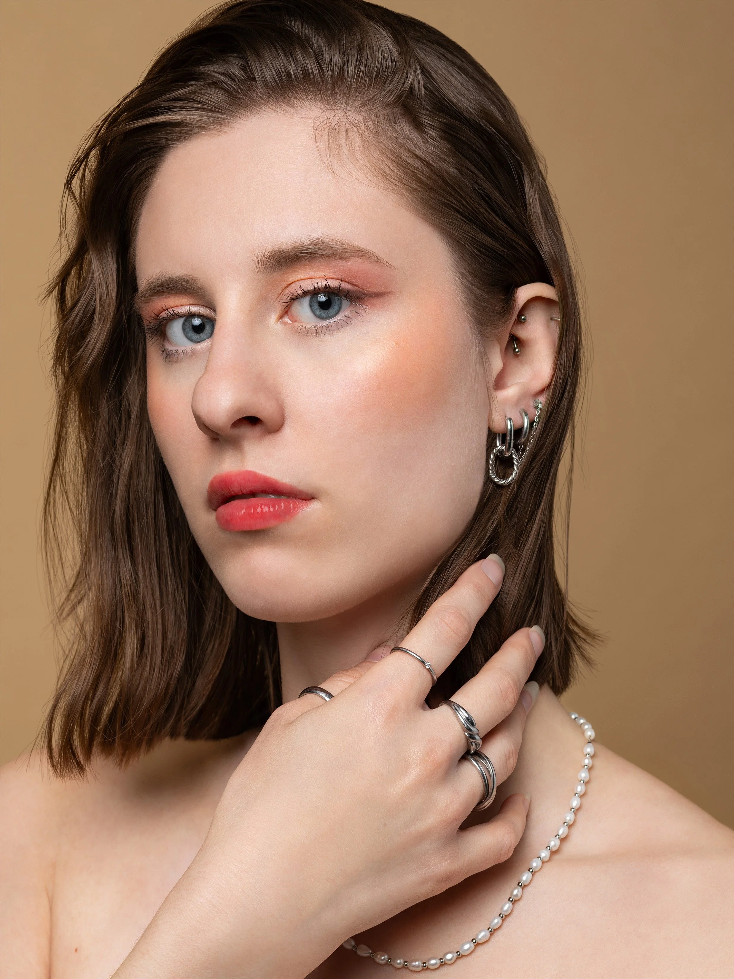 Close-up of a young woman with blue eyes and shoulder-length brown hair, wearing multiple silver rings, pearl necklace, and earrings, with a beige background.