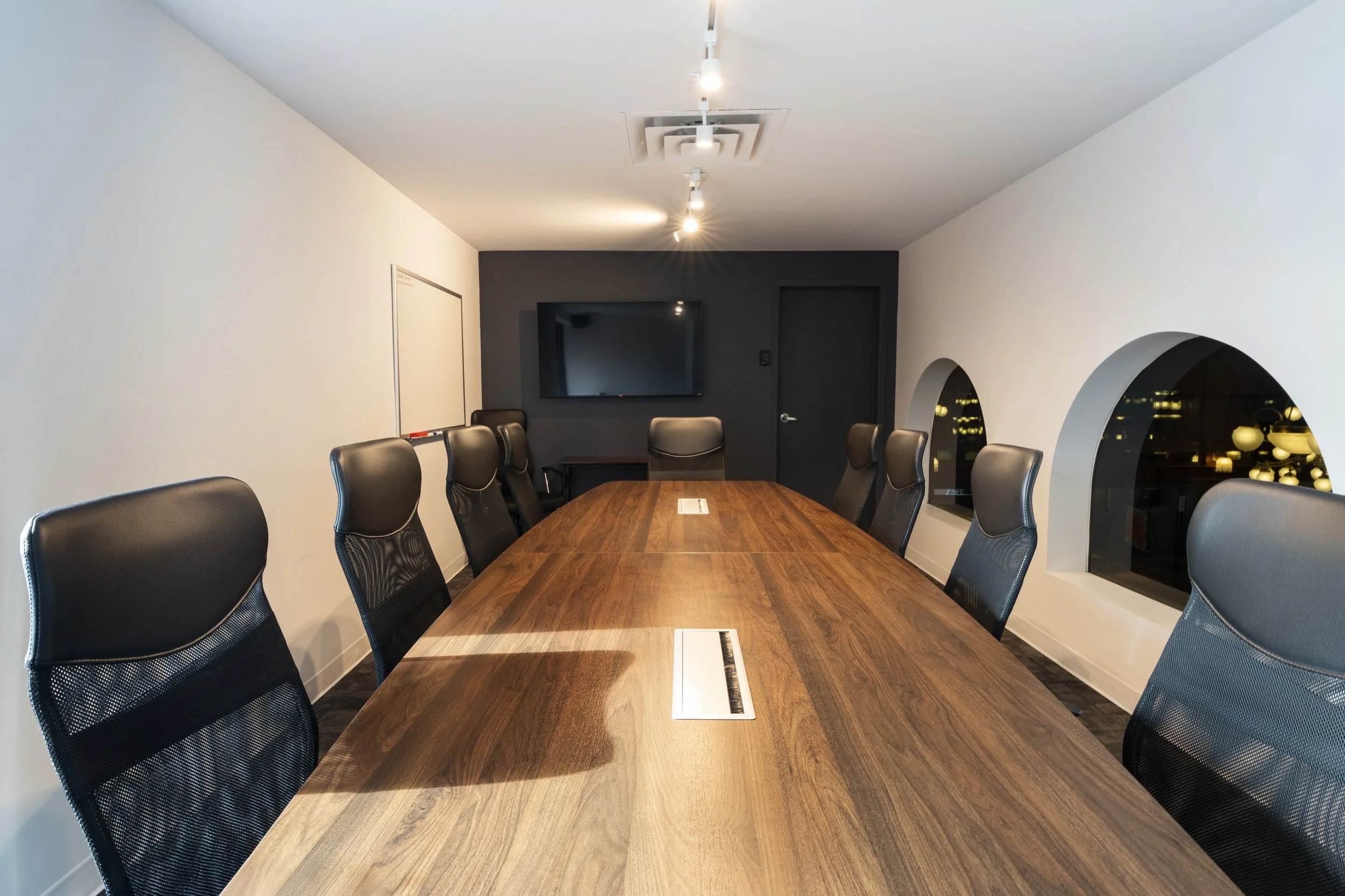 A modern conference room with a large wooden table and black office chairs. The room has white and dark gray walls, a wall-mounted flat-screen TV, and arched windows reflecting city lights at night.