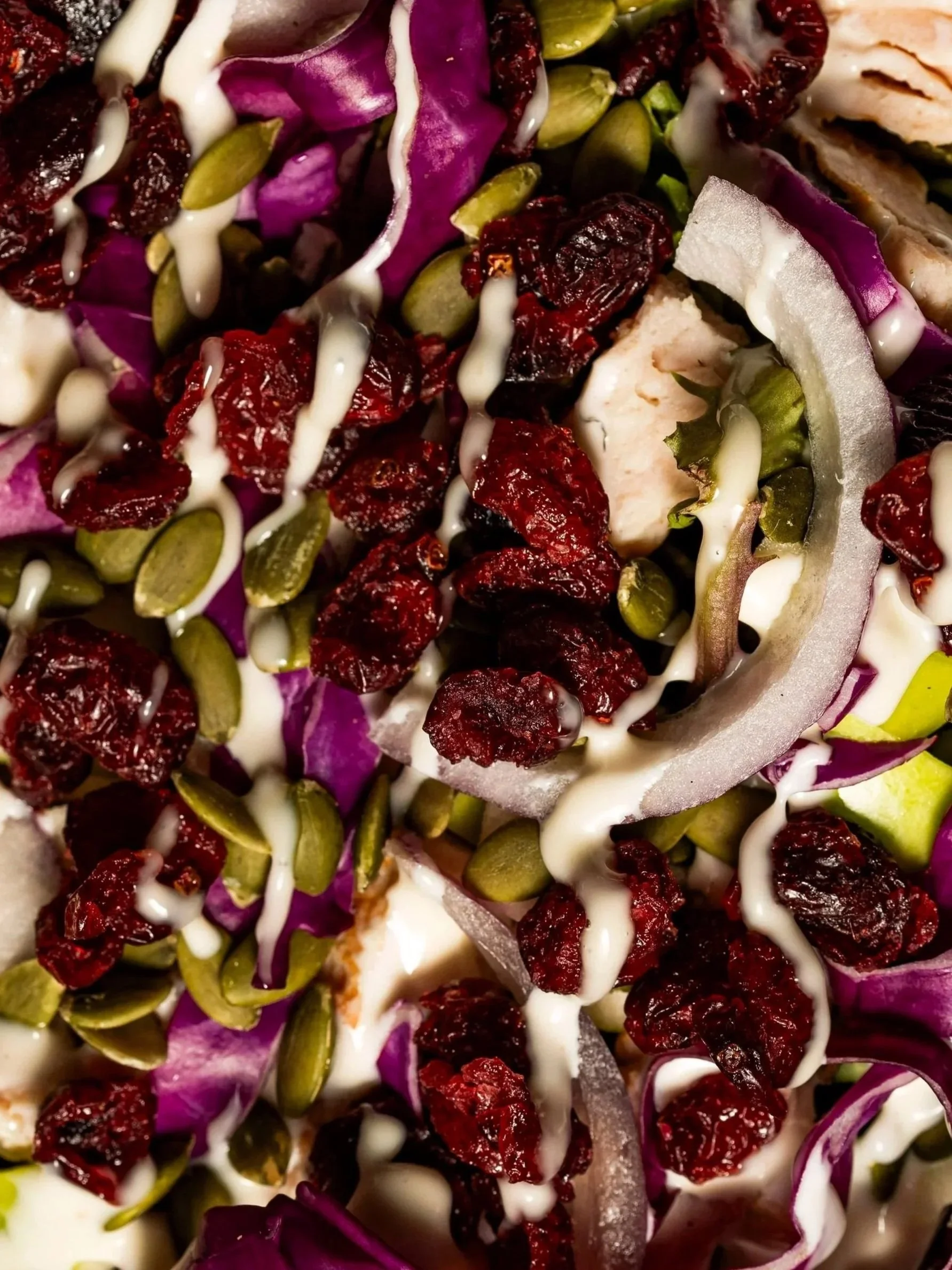 Close-up of a salad with red dried cranberries, green pumpkin seeds, purple cabbage, and a creamy white dressing drizzled on top.
