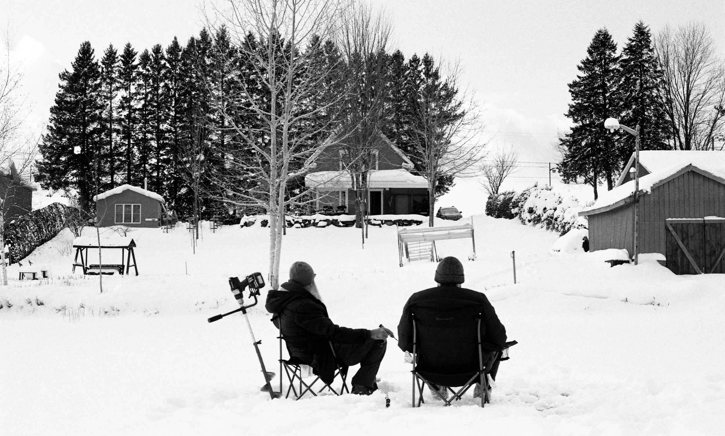 Two people sitting outdoors in a snowy backyard, facing away from the camera, with a tripod in front of them. The yard is covered in snow with trees, houses, and a fence in the background.