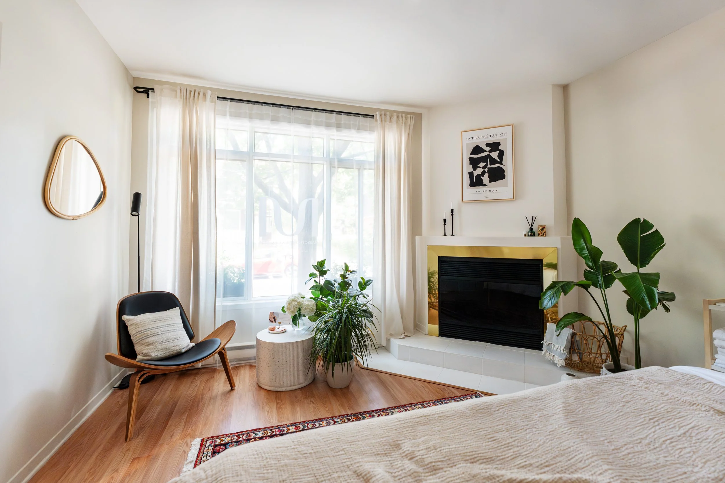 Living room with large window, beige curtains, green plants, framed artwork, black fireplace, armchair with pillow, small white table with flowers, eggs, and a book, hardwood flooring, and part of a bed in the foreground.