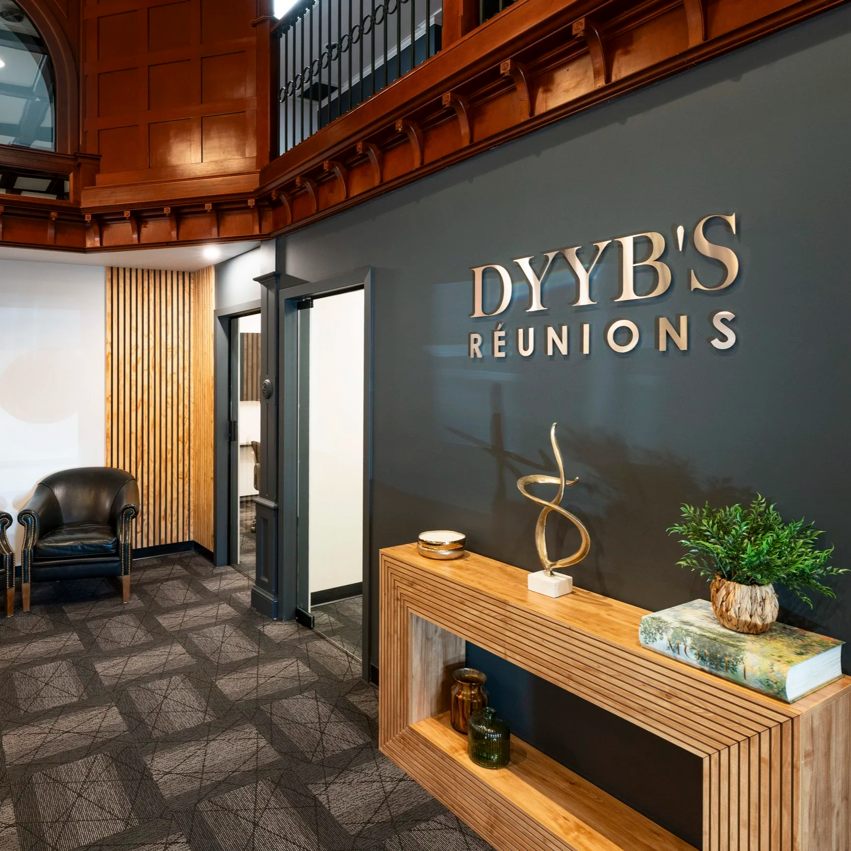 Reception area with a dark blue wall featuring the sign 'DYB'S RÉUNIONS,' a wooden table with decorative items including a large book titled 'MORFÉ' and sculptures, and a black leather armchair in the corner. The ceiling has wood paneling and railings with decorative black ironwork, and the room has a gray carpeted floor with a geometric pattern.