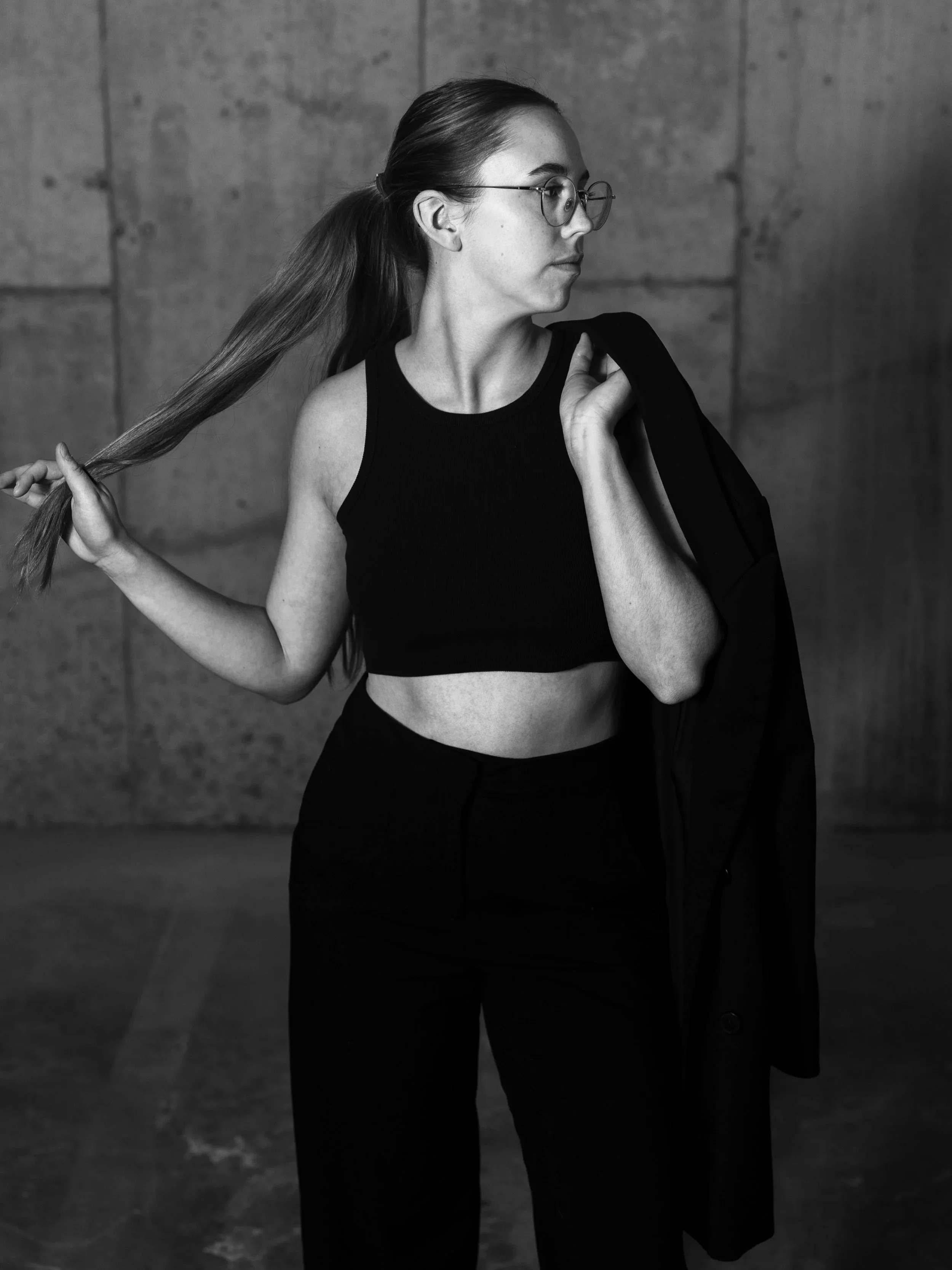 A woman in profile, wearing glasses and a black sleeveless top, holding a black jacket over her shoulder, standing against a concrete wall background in a black-and-white photograph.