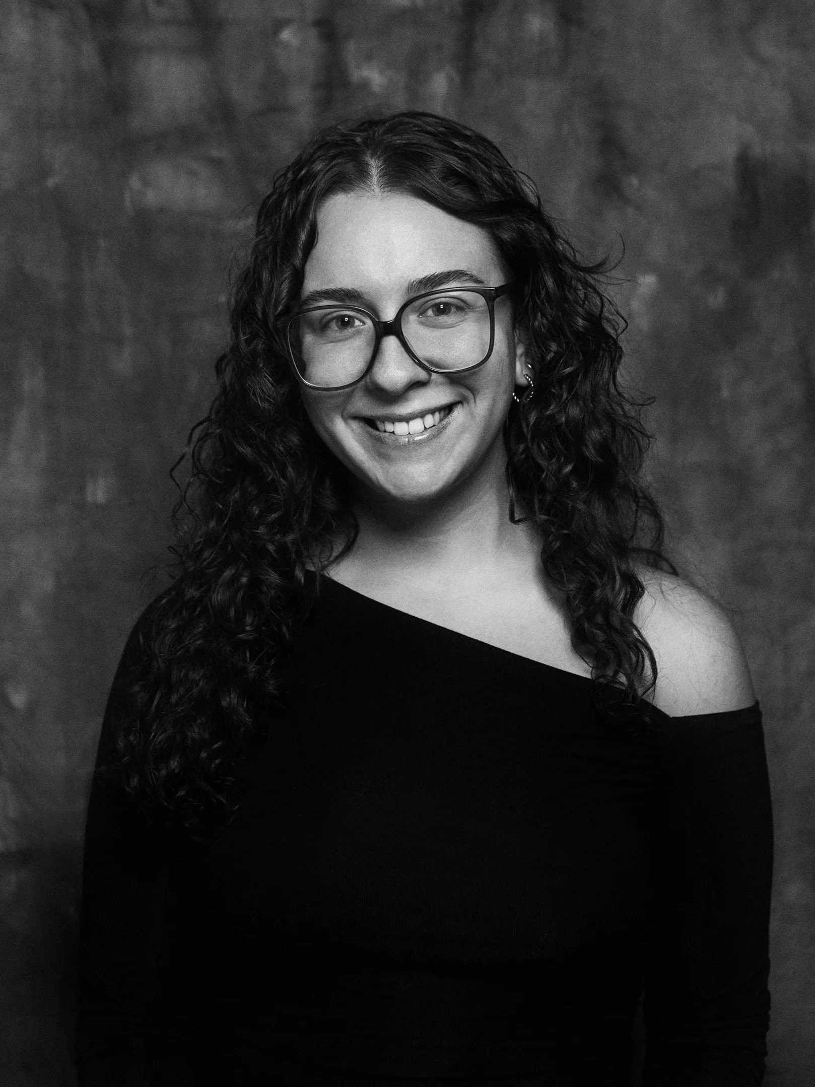 Black and white portrait of a smiling woman with curly hair, wearing large glasses and a one-shoulder top, against a textured background.