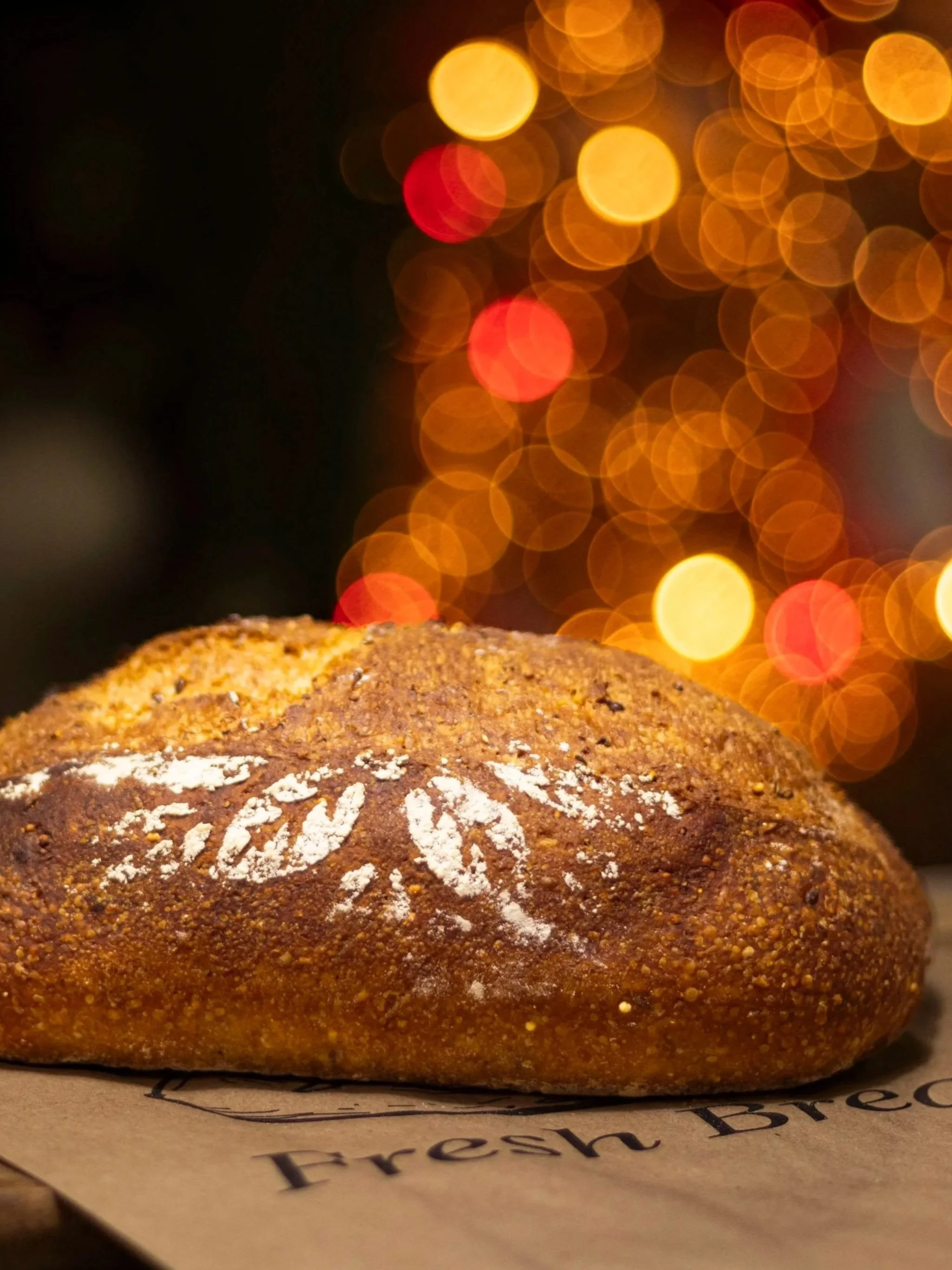 Close-up of a baked bread with a golden-brown crust, sprinkled with white flour, against a backdrop of blurred warm-colored lights.