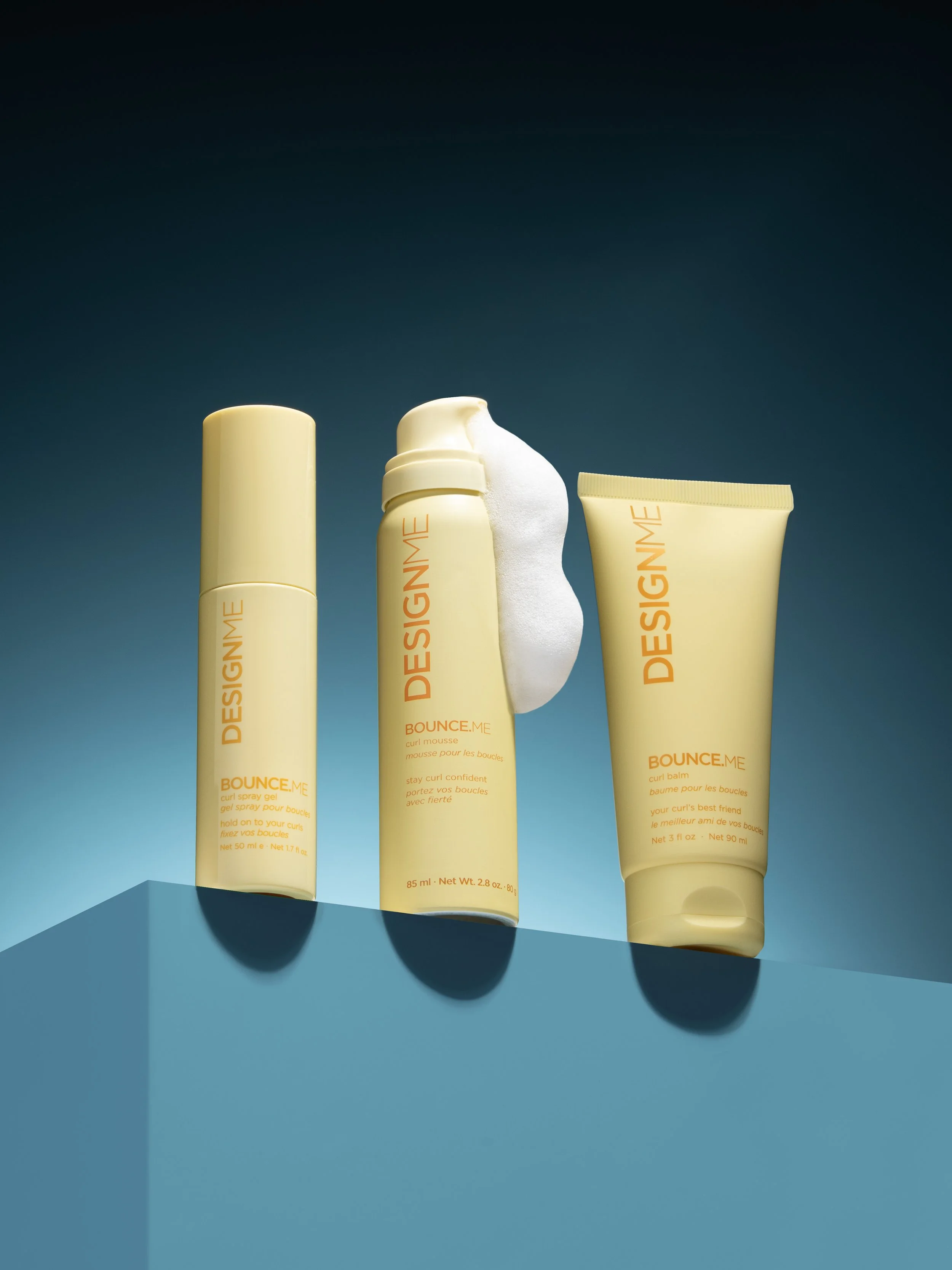 Three yellow cosmetic containers on a blue surface with a dark blue background. The containers are labeled with orange text including "DESIGNME" and product names, one with foam coming out of its opening.