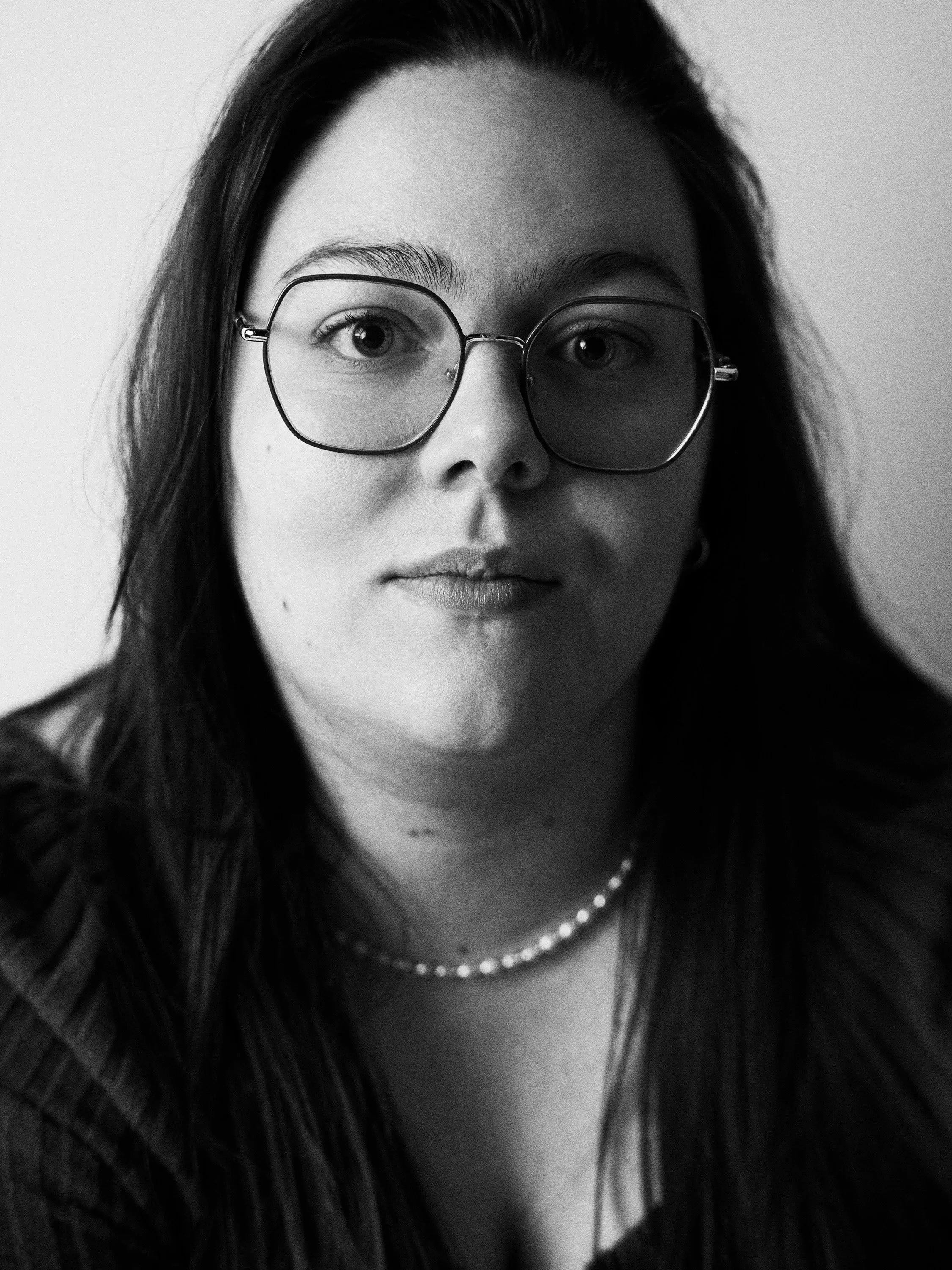 Black and white portrait of a woman wearing glasses, with long hair, a pearl necklace, and a pulled-up top.