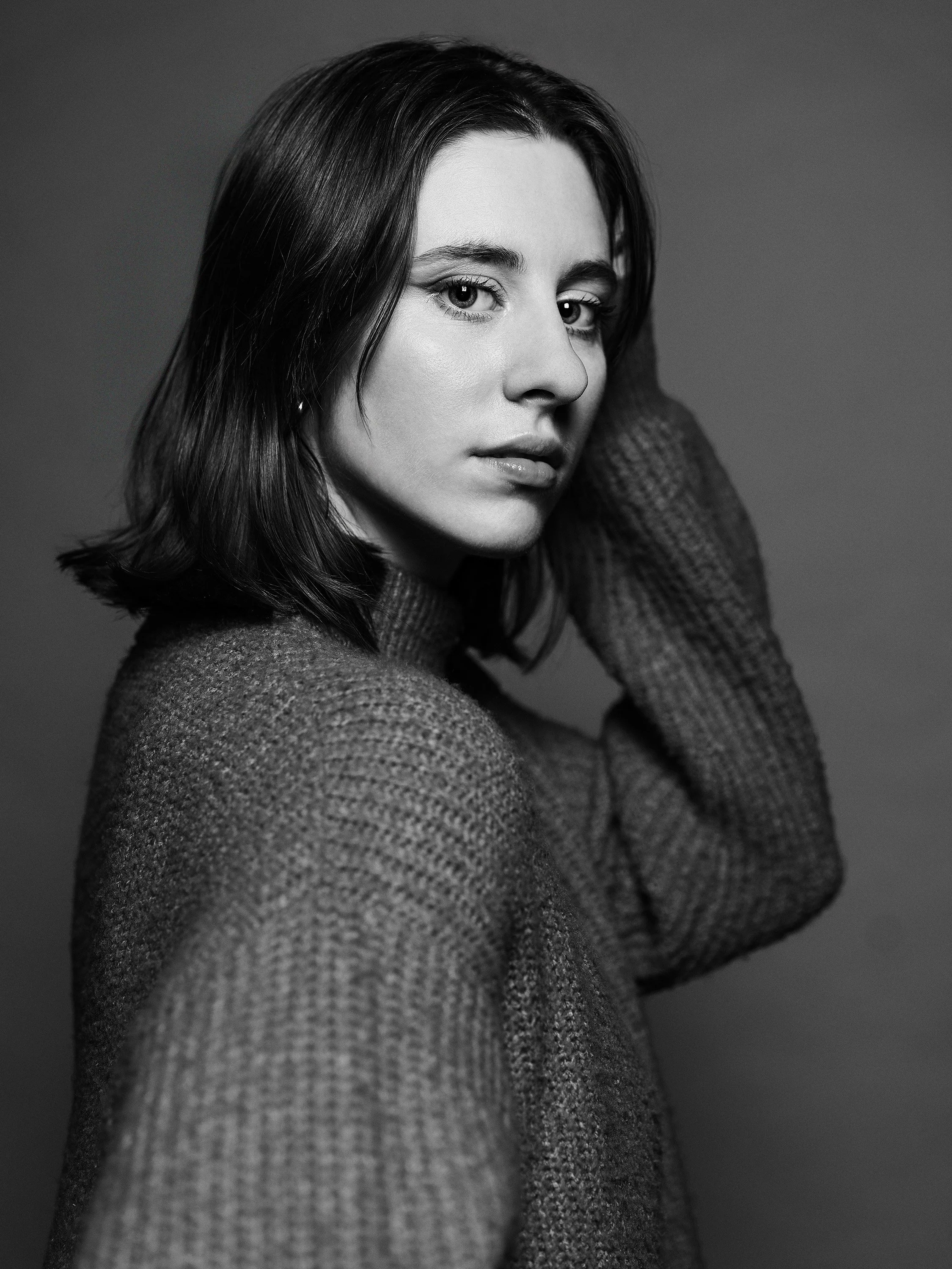 Black and white portrait of a woman with shoulder-length hair, wearing a knitted sweater, with one hand near her face, looking directly at the camera.