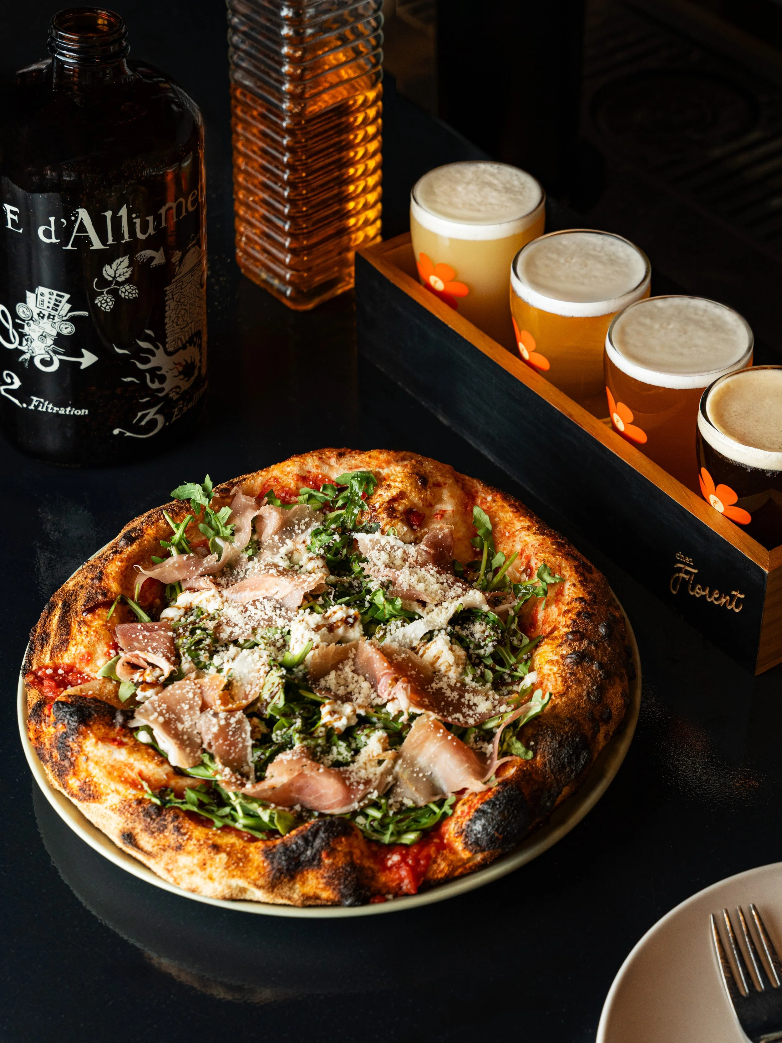 Close-up of a pizza with arugula, shredded cheese, and prosciutto on a white plate. In the background, there's a dark table with a bottle labeled 'Eau d'Allume' and a wooden box holding four glasses of beer with orange flower designs, and a glass bottle with a textured surface.