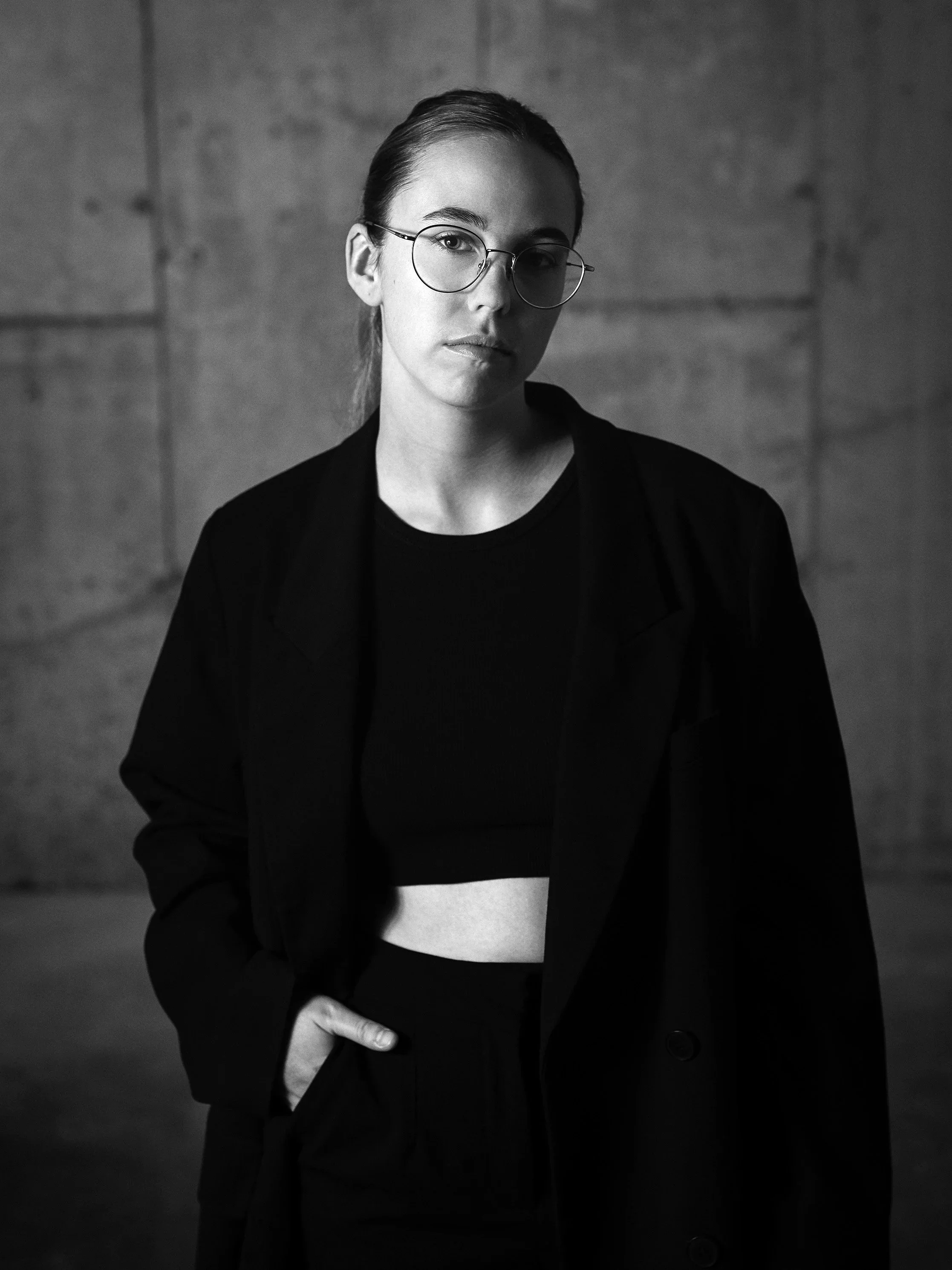 A black and white photo of a woman with glasses, wearing a black blazer over a cropped top, standing against a plain wall. She has her hair pulled back and is looking directly at the camera with a neutral expression.