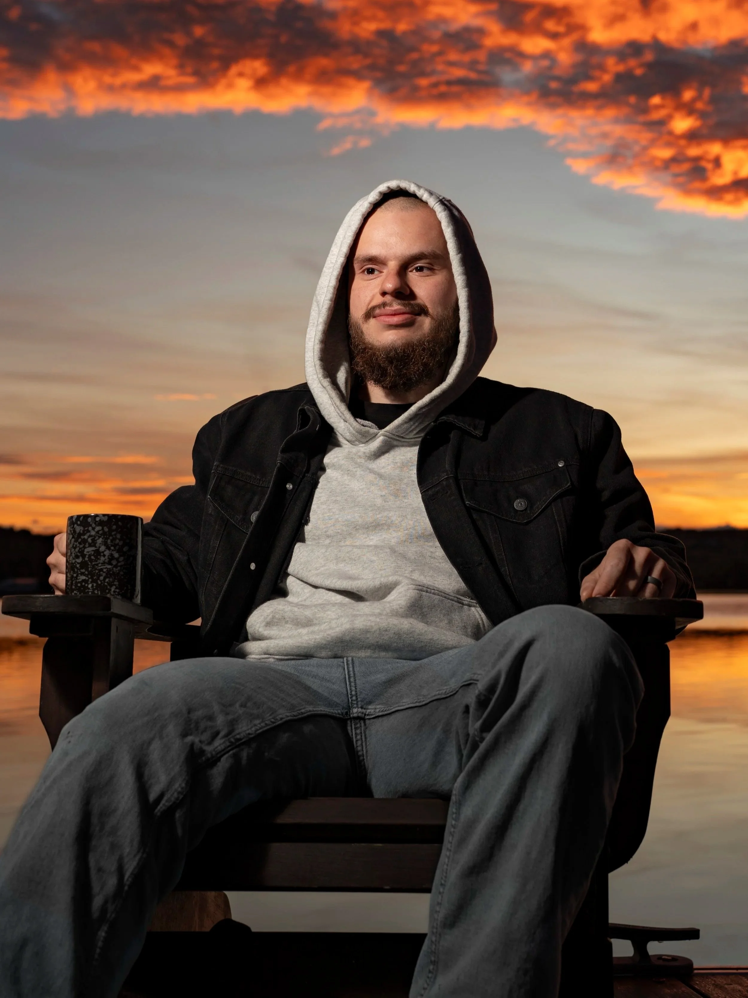A man with a beard and hoodie sitting outdoors at sunset, with a colorful sky and clouds in the background.