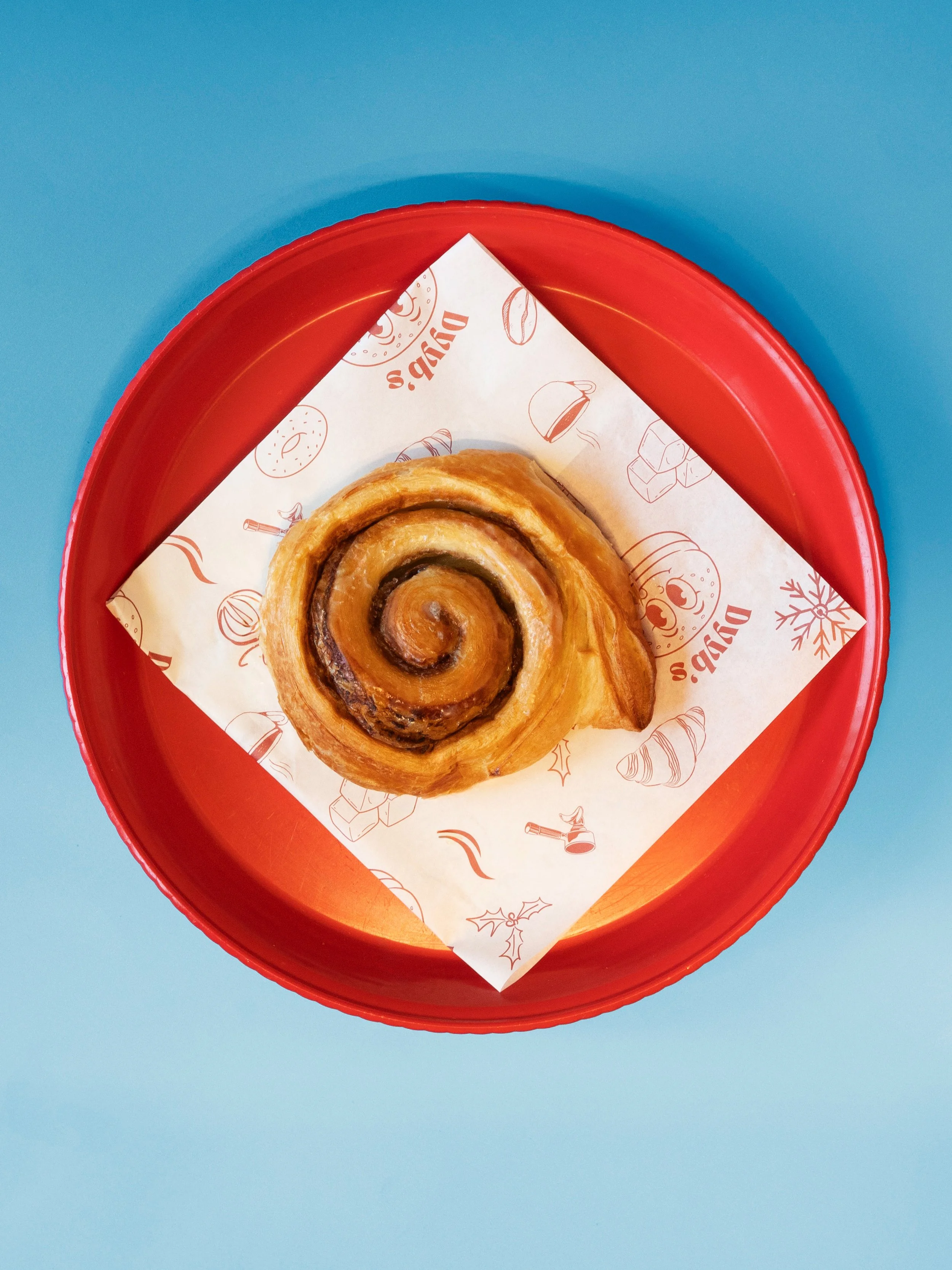Close-up of cinnamon roll on red tray with holiday themed paper, against a blue background.