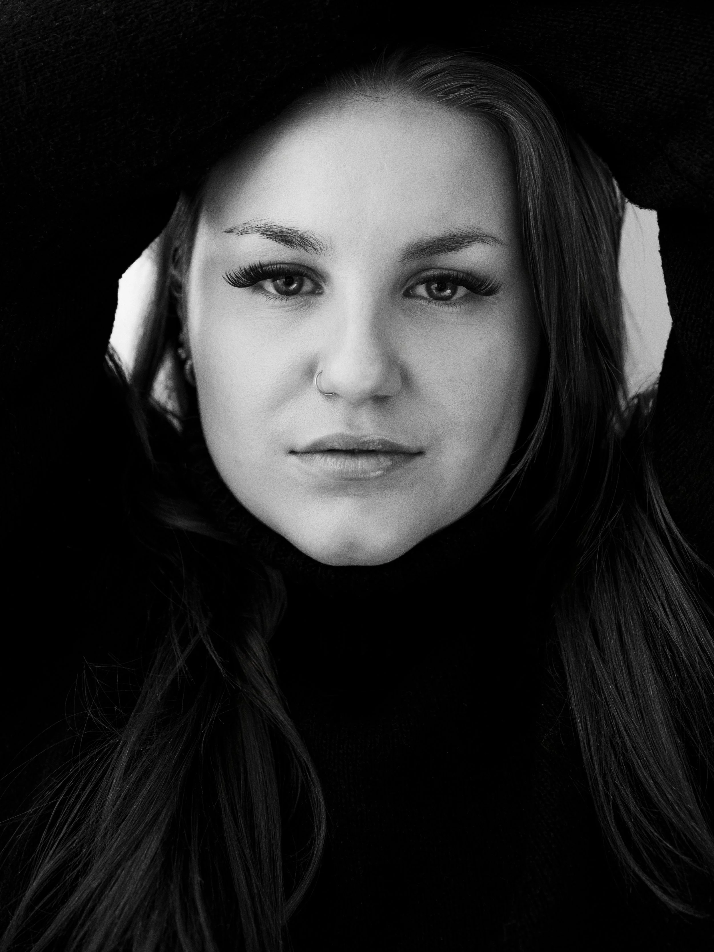 Black and white portrait of a woman with long hair, looking directly at the camera, with one eyebrow slightly raised and a nose piercing.