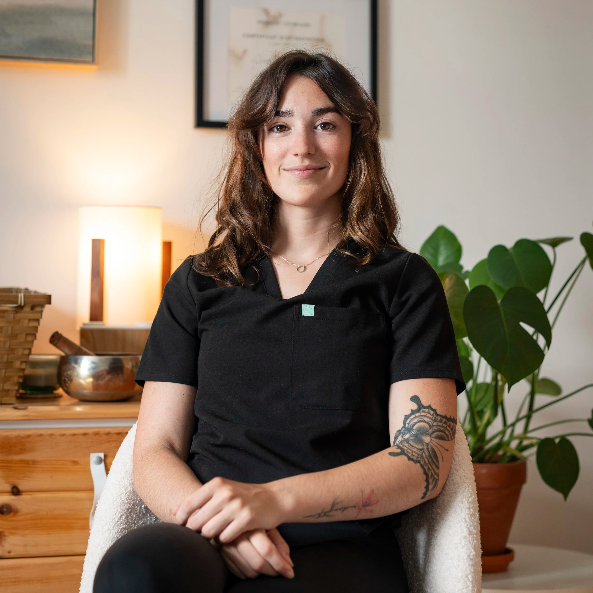 A woman with wavy brown hair and tattoos on her arm, smiling and sitting indoors in a room with a wooden dresser, a potted plant, and framed artwork on the wall.
