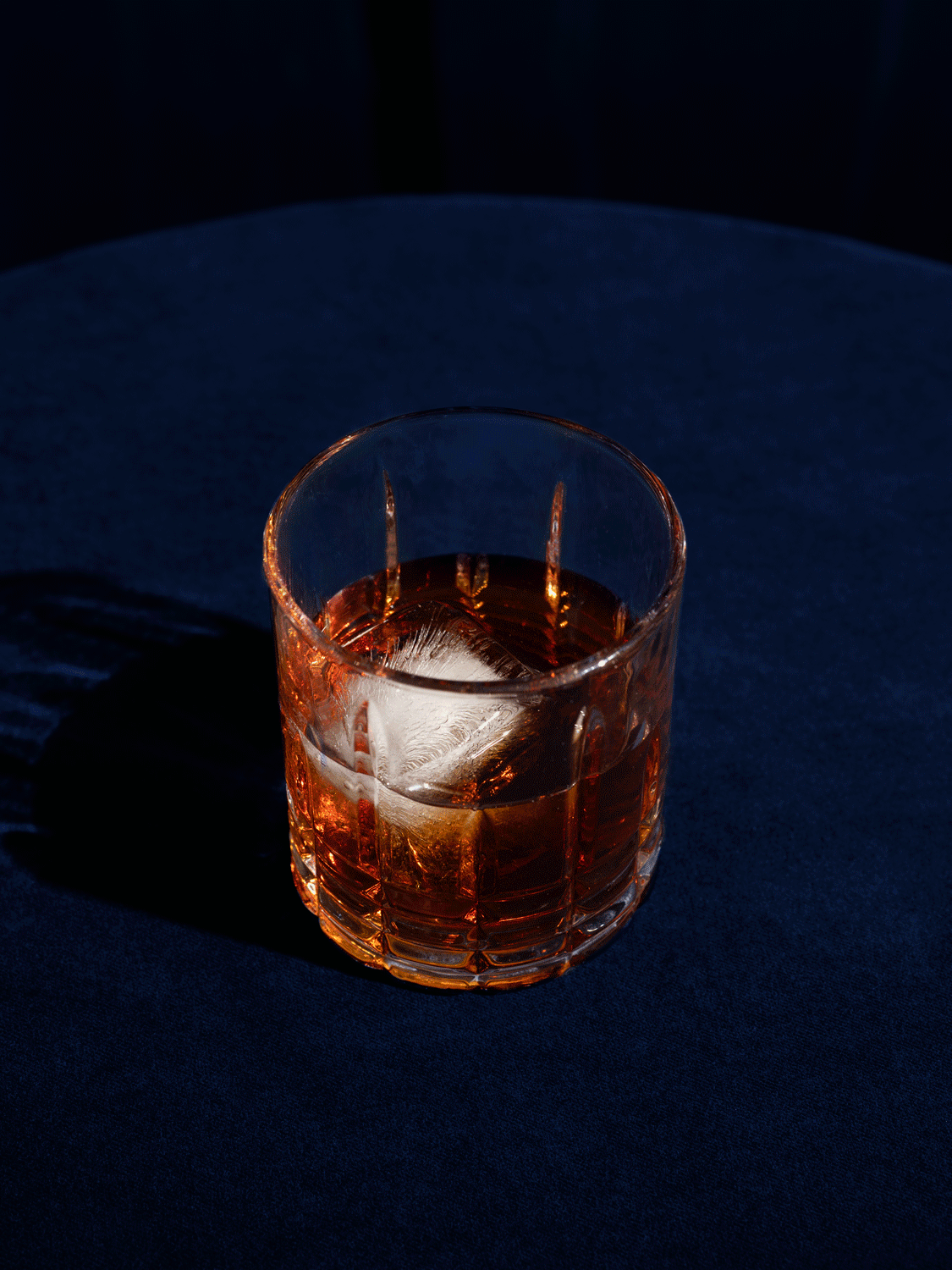 A glass of whiskey with a large ice cube on a dark blue surface.