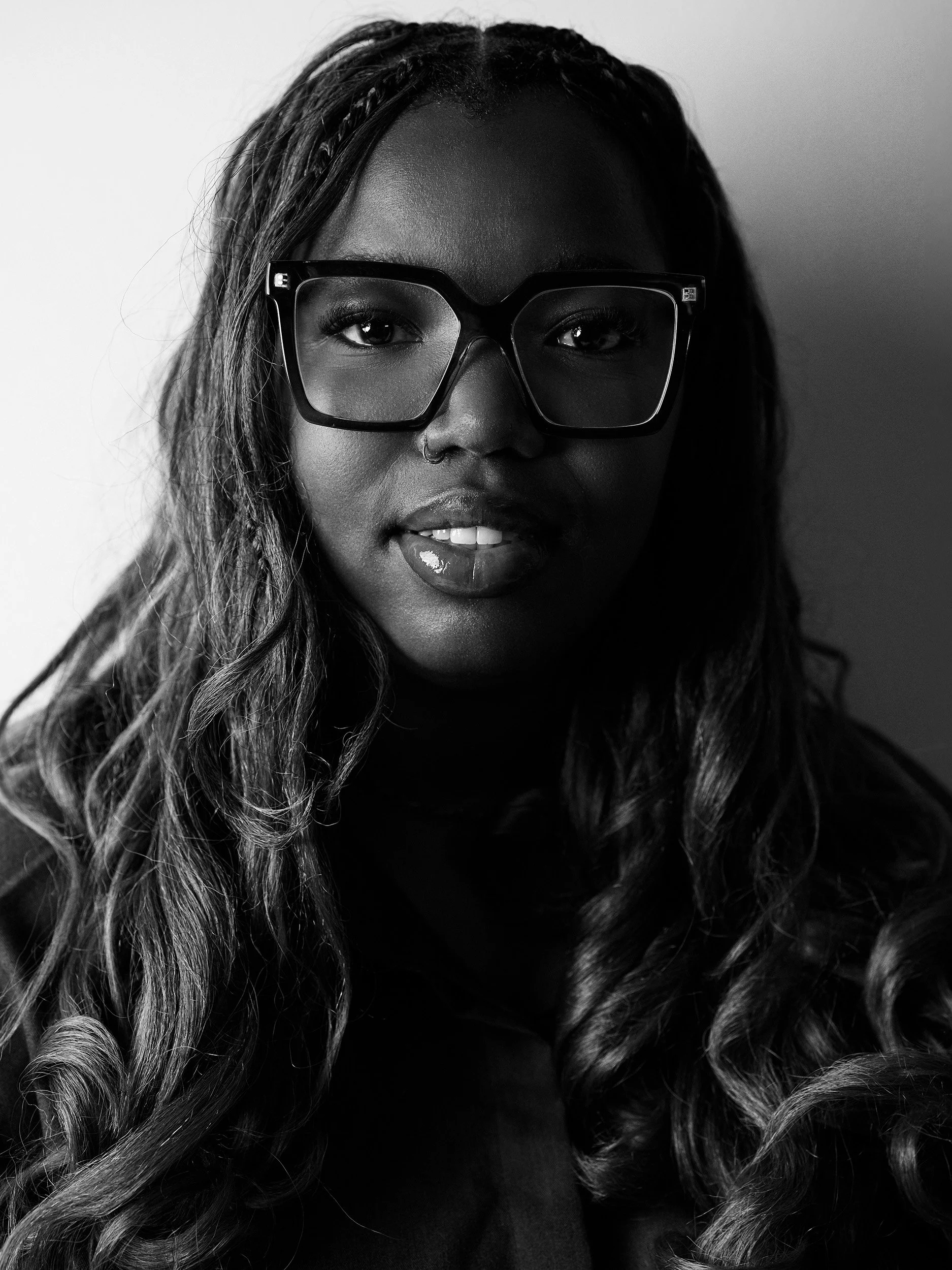 A black-and-white portrait of a woman with long, wavy hair wearing large, square-shaped glasses, and a nose ring. She is looking directly at the camera.