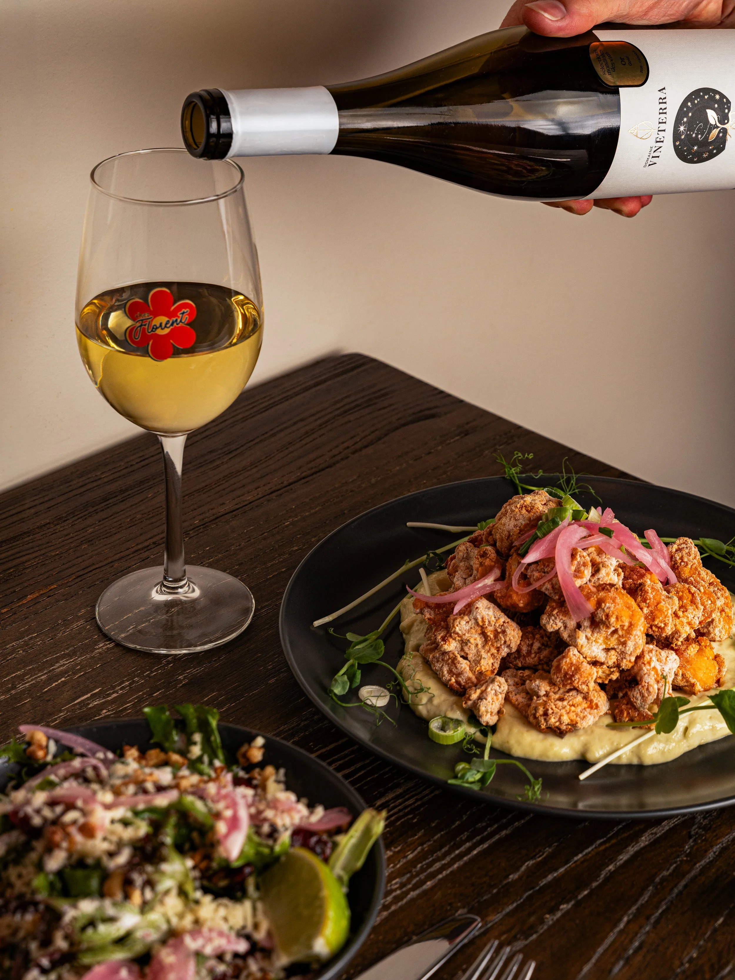 A hand pouring white wine from a bottle into a wine glass on a wooden table, with a plate of fried chicken topped with pickled onions and garnishes nearby.