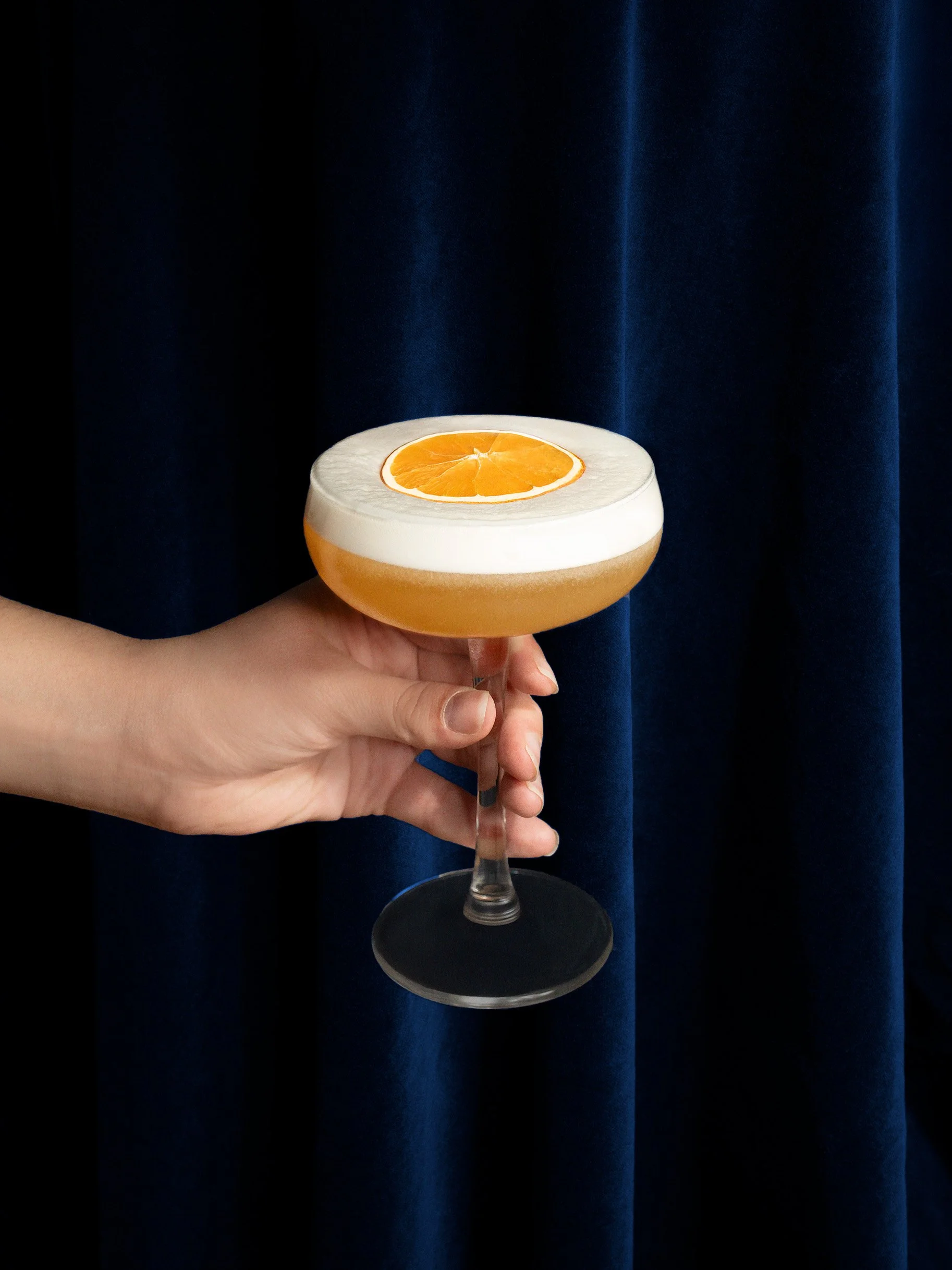 A hand holding a cocktail glass with a layer of white foam and a slice of orange on top, set against a dark blue curtain background.