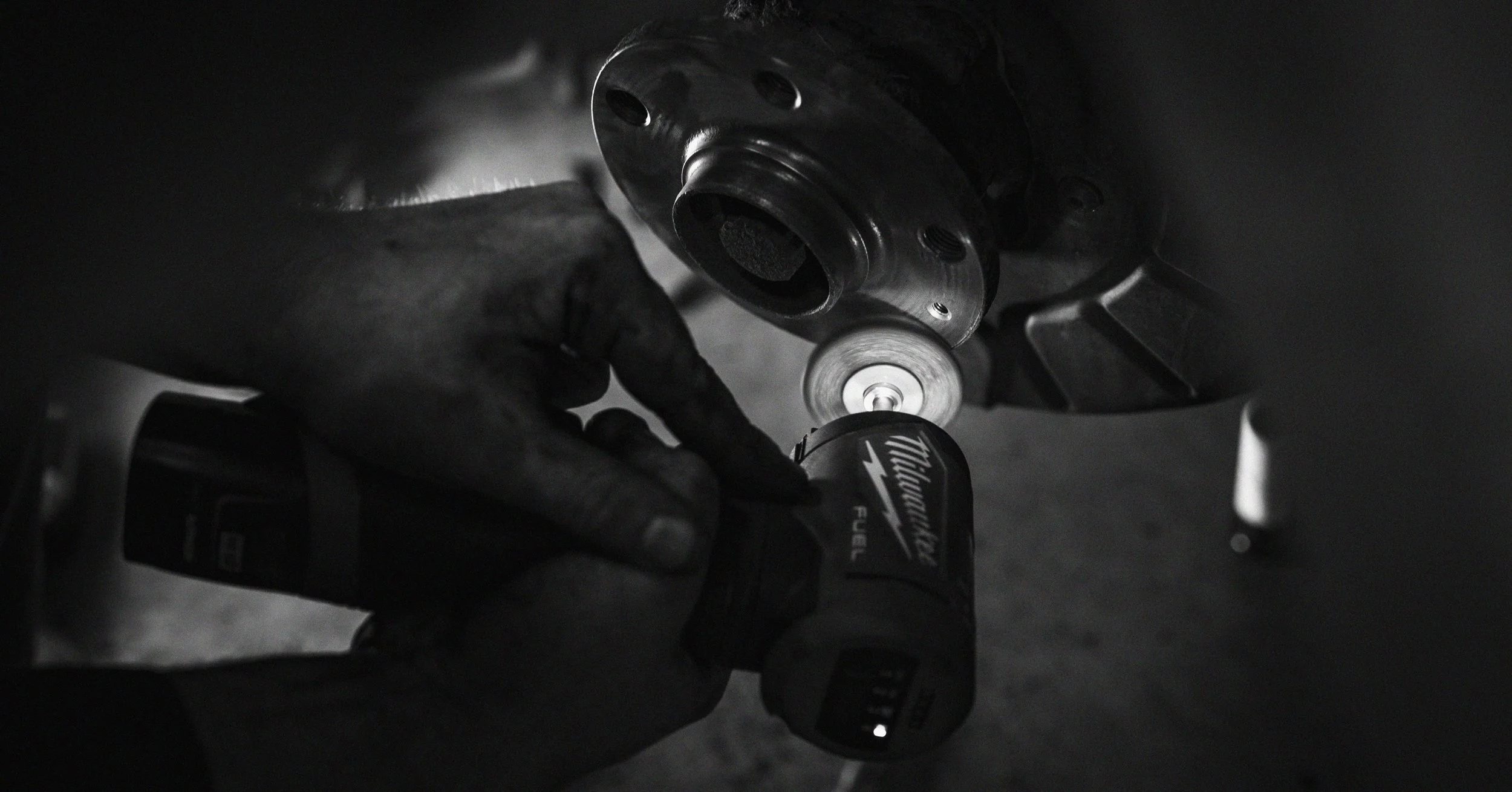 A person wearing black gloves is using a cordless drill to work on a metal vehicle part in a dark environment.