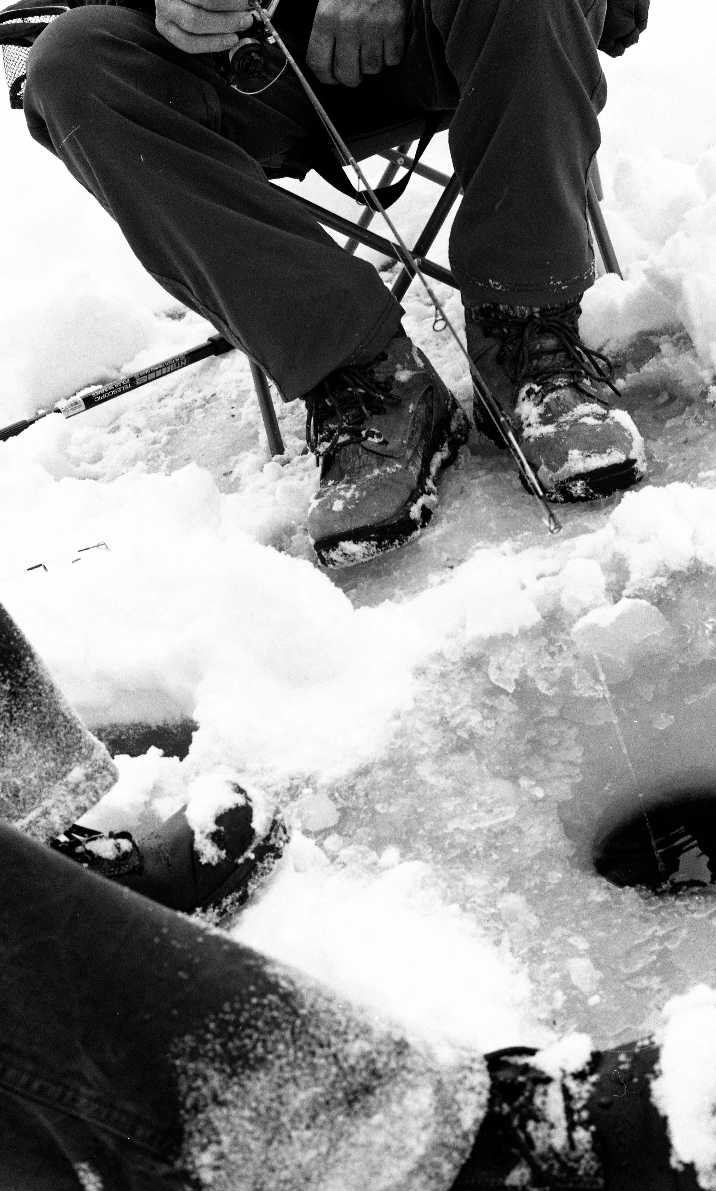 Person sitting in a folding chair on snow-covered ground, wearing winter clothing and sturdy boots, with fishing gear nearby.