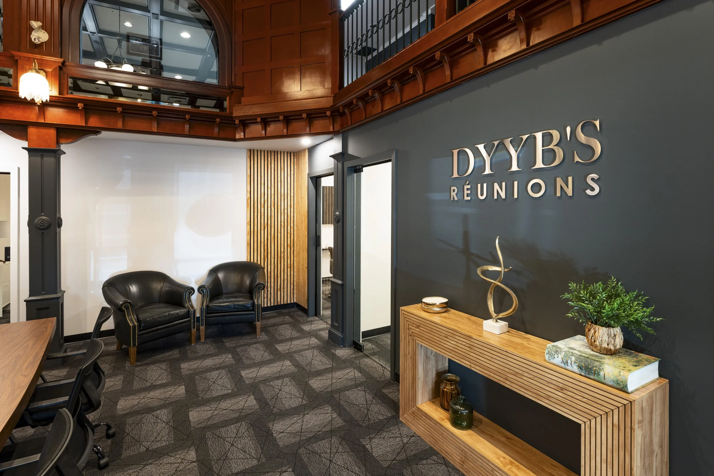 Reception area with a dark blue wall featuring the sign 'DYB'S RÉUNIONS,' a wooden table with decorative items including a large book titled 'MORFÉ' and sculptures, and a black leather armchair in the corner. The ceiling has wood paneling and railings with decorative black ironwork, and the room has a gray carpeted floor with a geometric pattern.