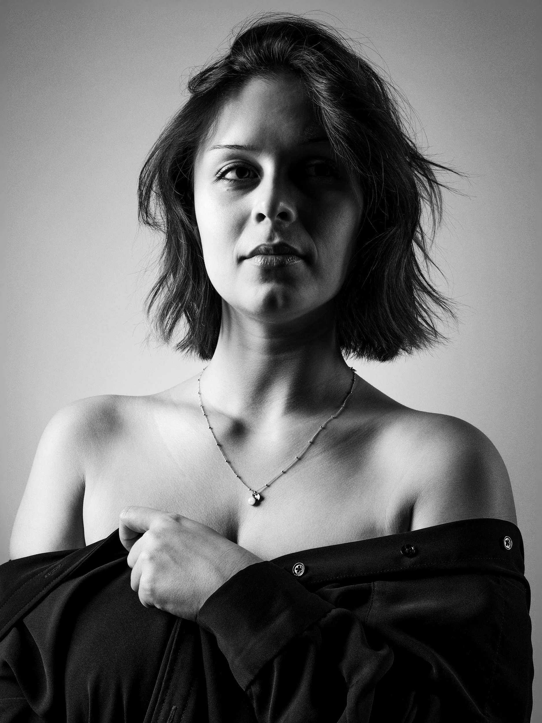 Black and white photograph of a woman with shoulder-length slightly wavy hair, wearing a necklace and partially removing a black jacket, with a neutral background.