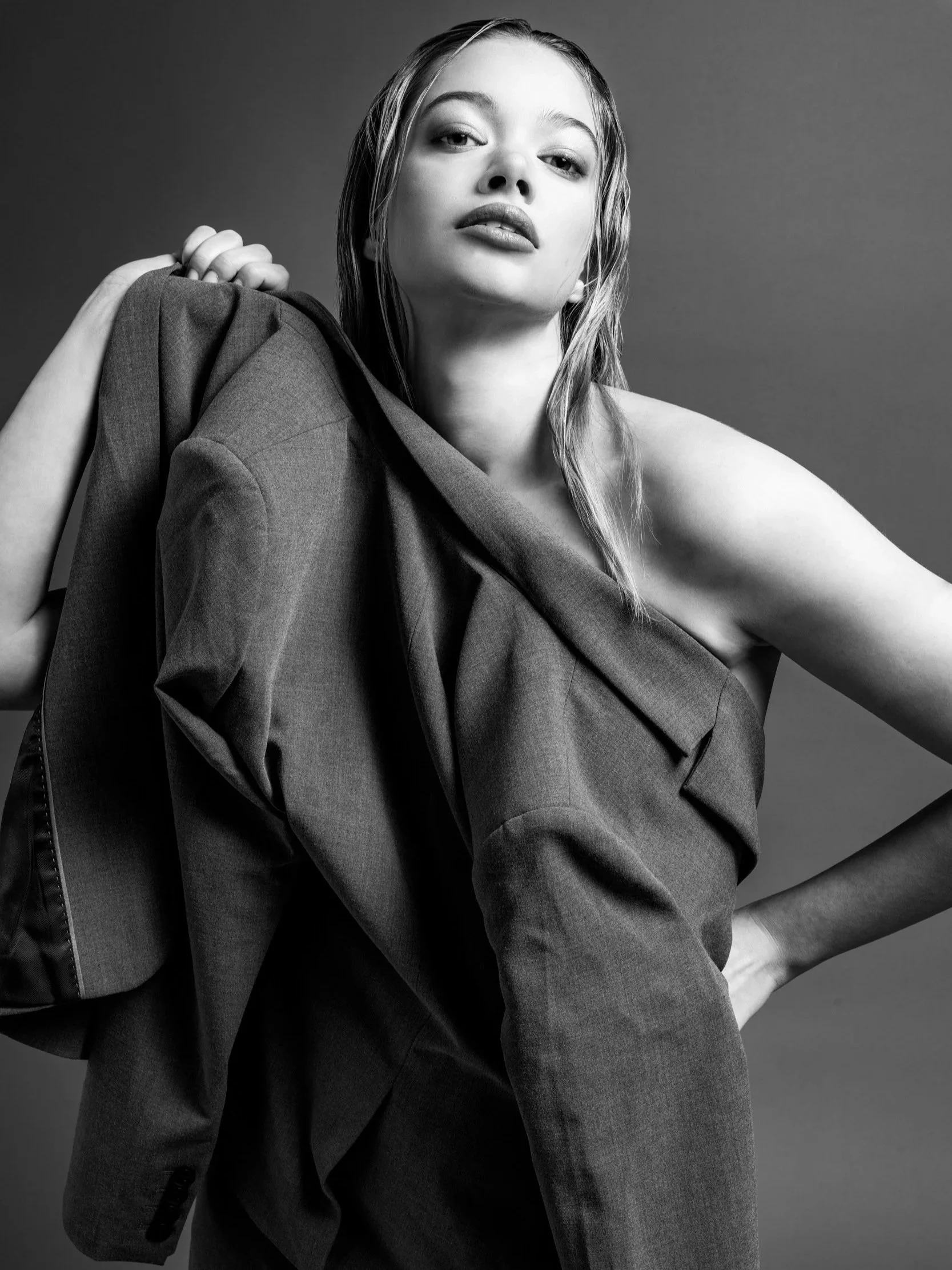 Black and white photo of a woman with wet hair, wearing a jacket draped over her shoulder, standing against a plain background.