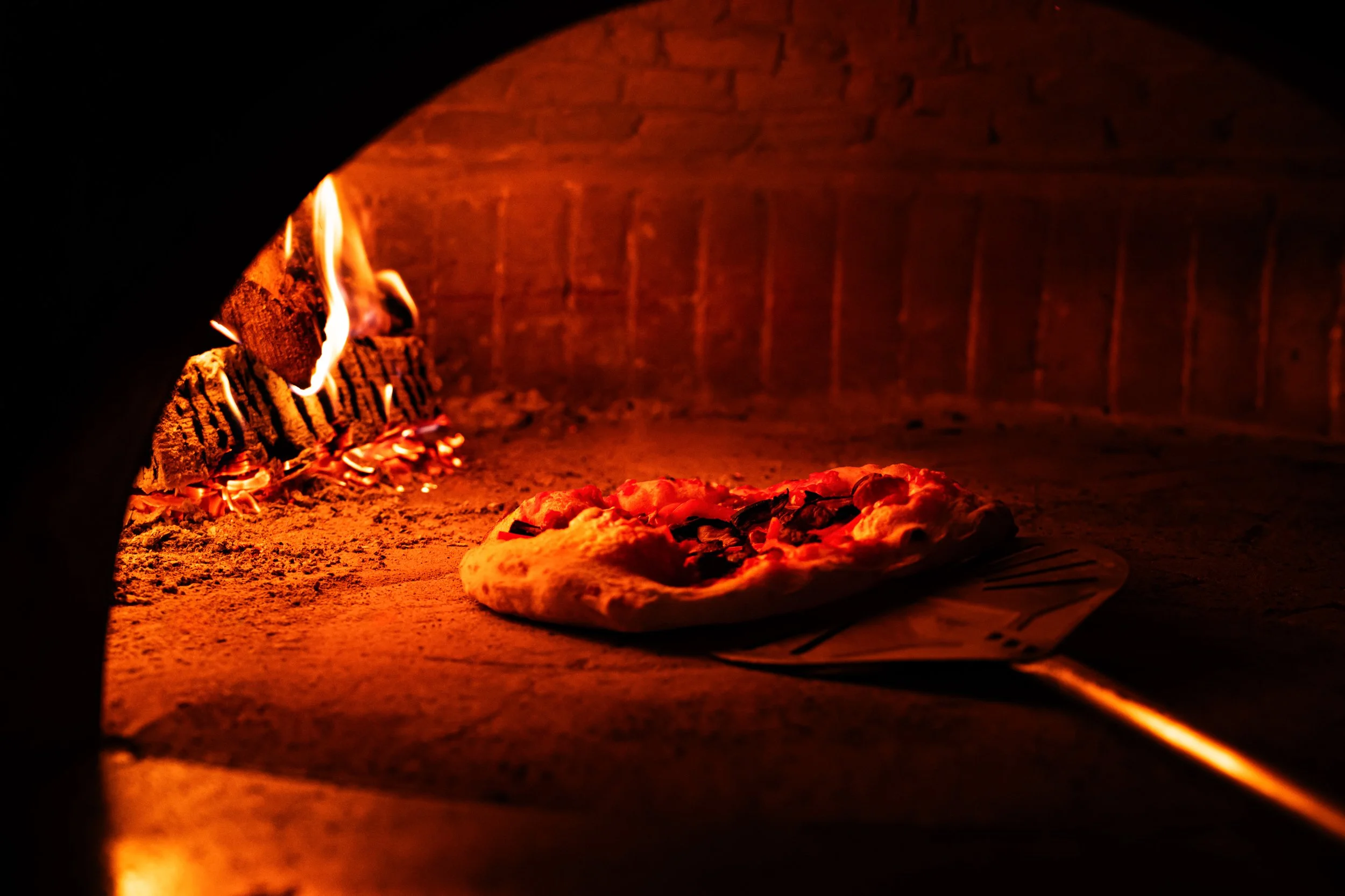 Pizza baking inside a wood-fired oven with glowing flames and a brick interior.